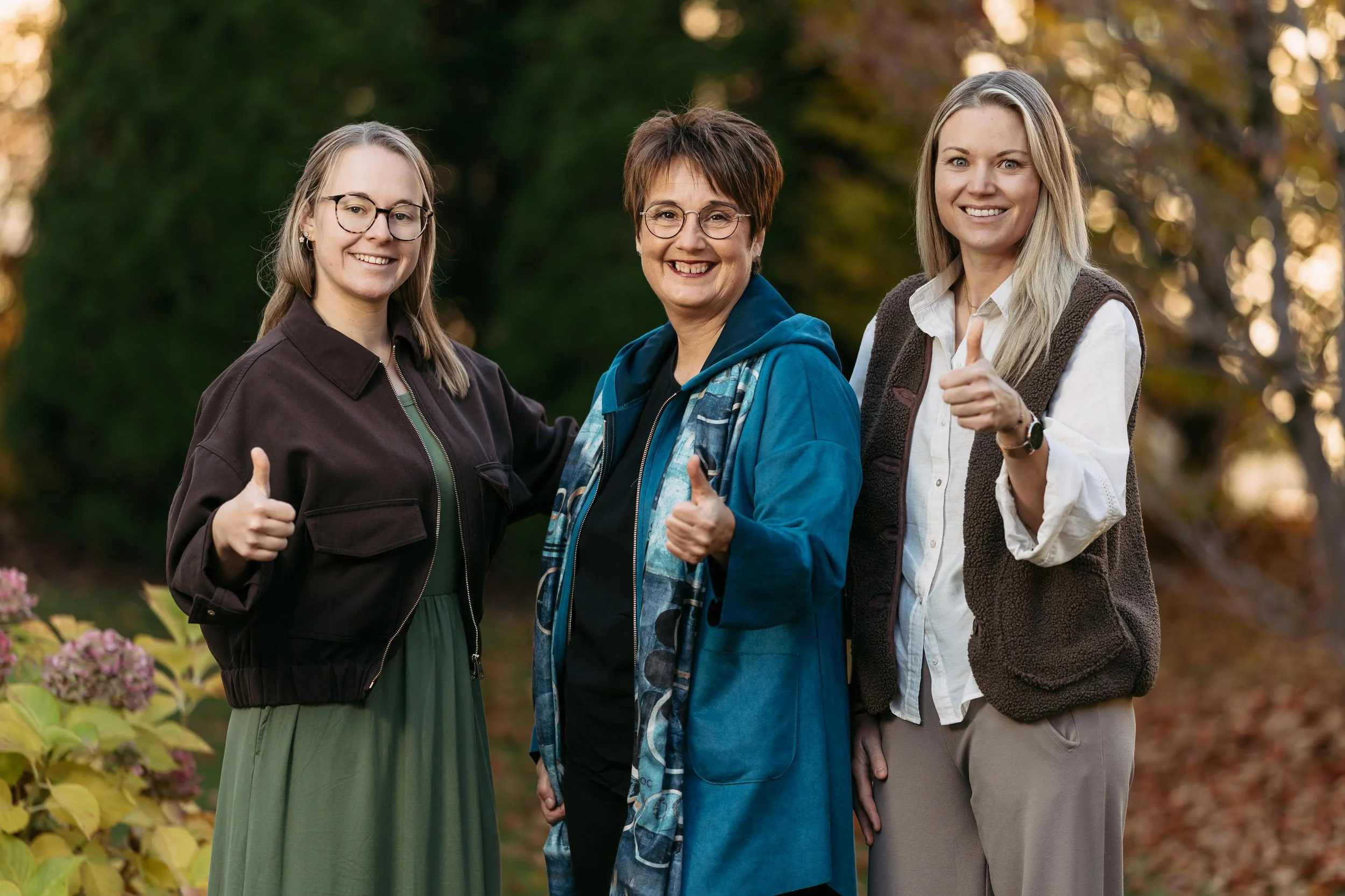 Drei Frauen stehen im Park im Herbst, lachen und machen Daumen hoch.