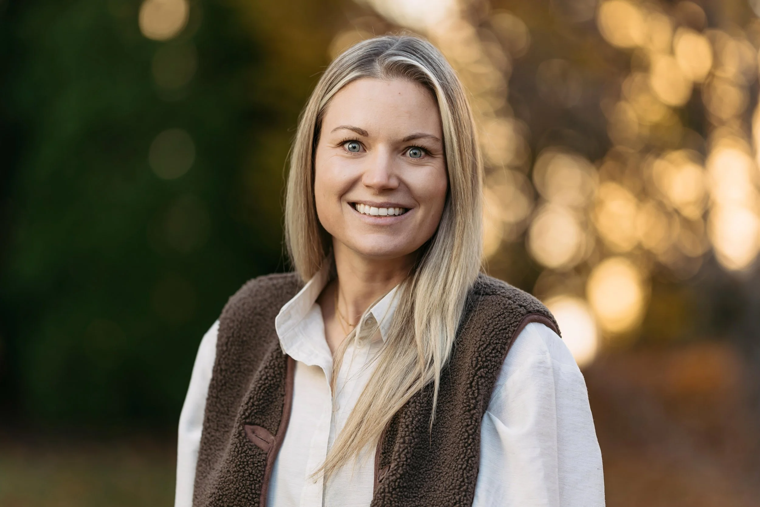 Lächelnde blonde Frau im Freien mit bunter Herbstkulisse im Hintergrund.