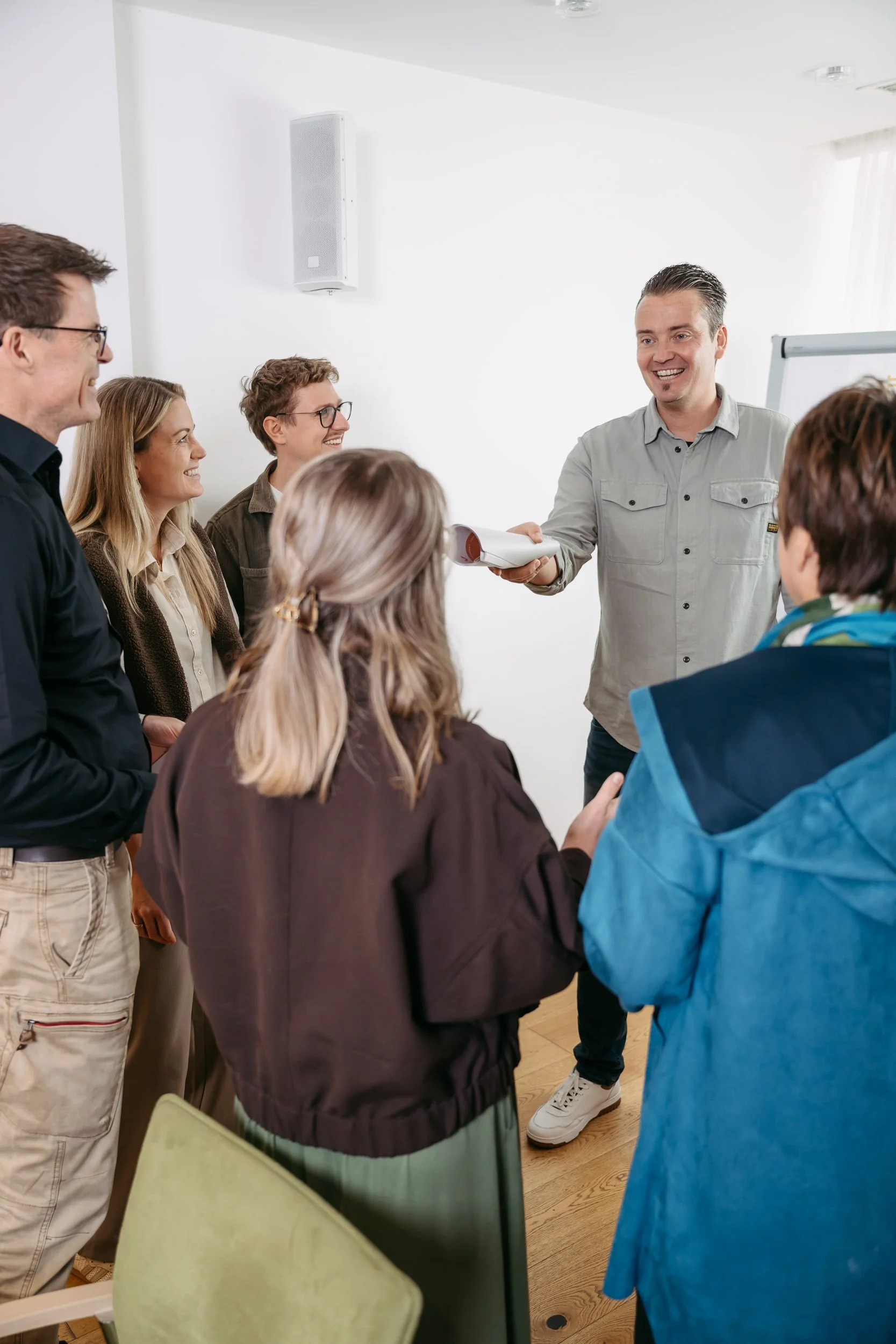 Ein Mann übergibt einem anderen Mann ein Dokument während einer Präsentation vor einer Gruppe von Menschen in einem hellen Raum.