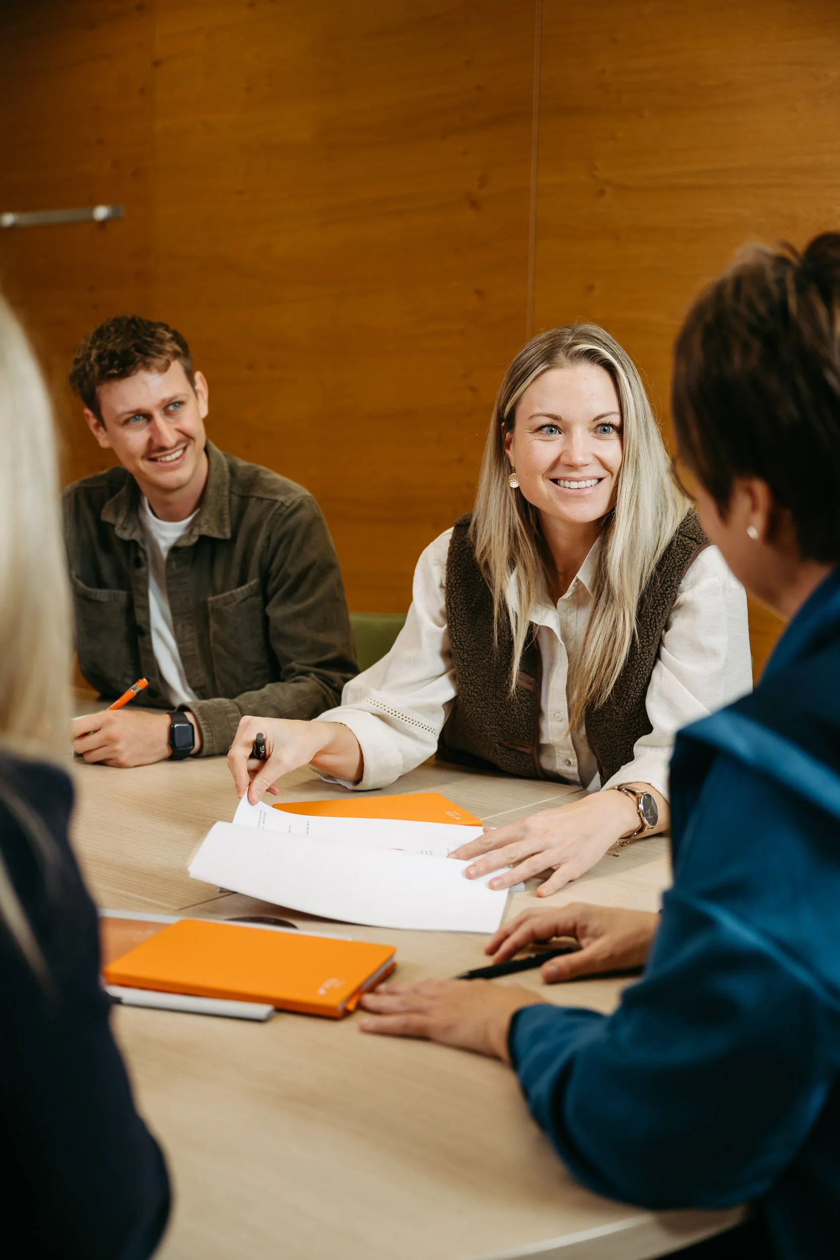 Vier Personen sitzen um einen Tisch in einem Gespräch, einige haben Notizbücher und Stifte, und sie sind in einem Raum mit Holzvertäfelung im Hintergrund.