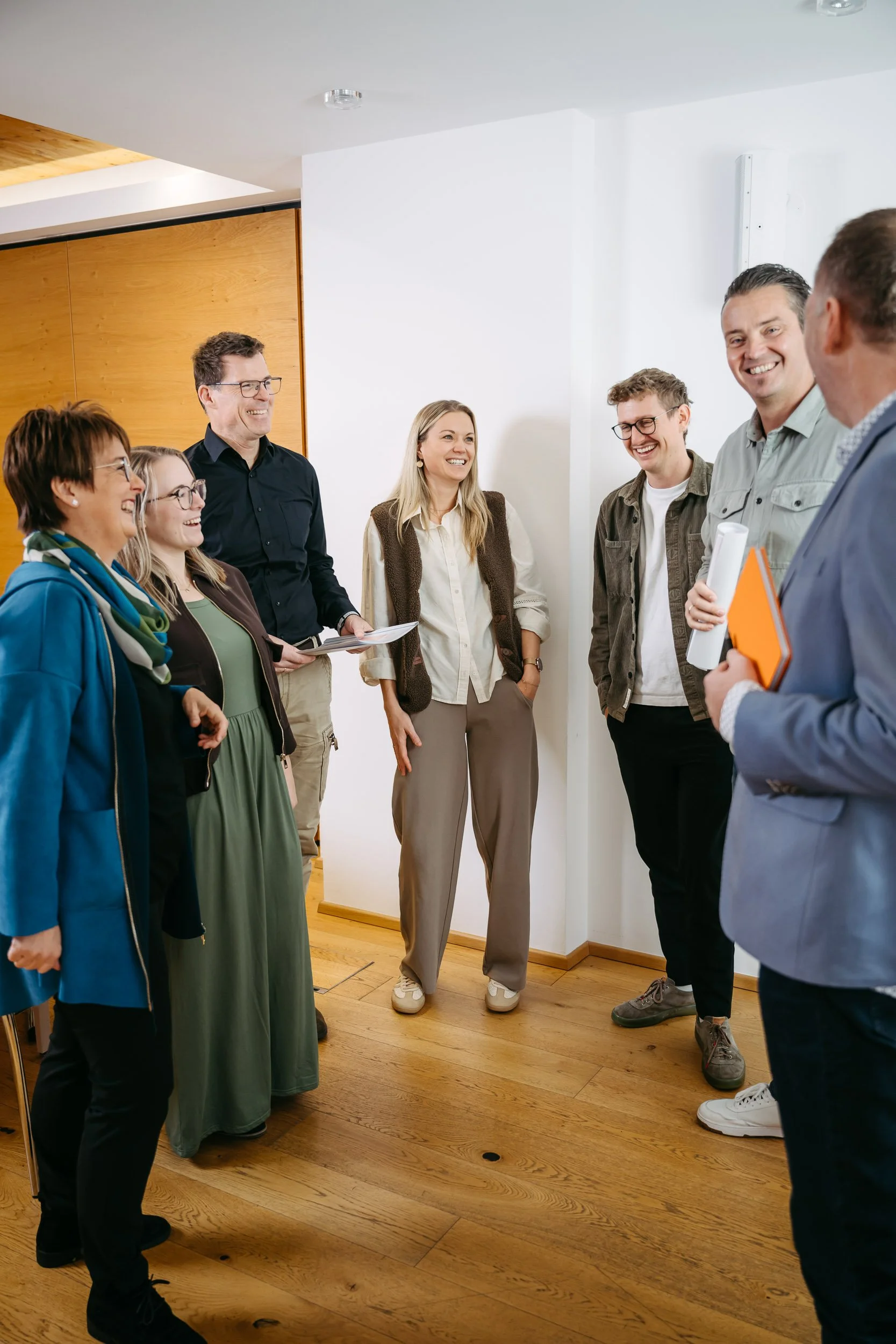 Menschen stehen in einem Büro und lachen gemeinsam in einer Gruppe, einige halten Dokumente.»