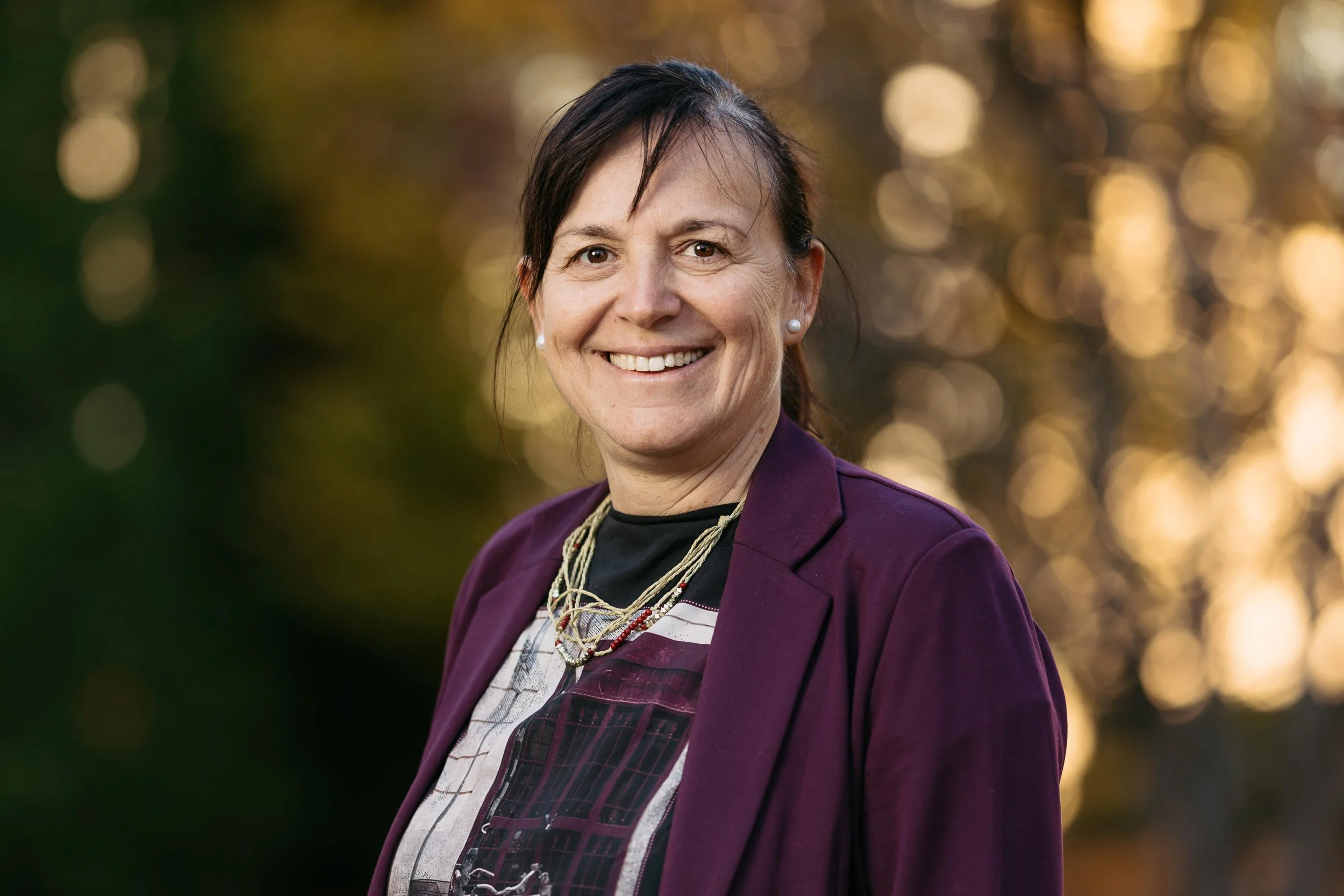 Eine lachende Frau mit dunklem Haar, die in einem lila Blazer und einer schwarzen Bluse mit grafischem Muster steht, vor einem herbstlichen Hintergrund mit goldenem Licht.