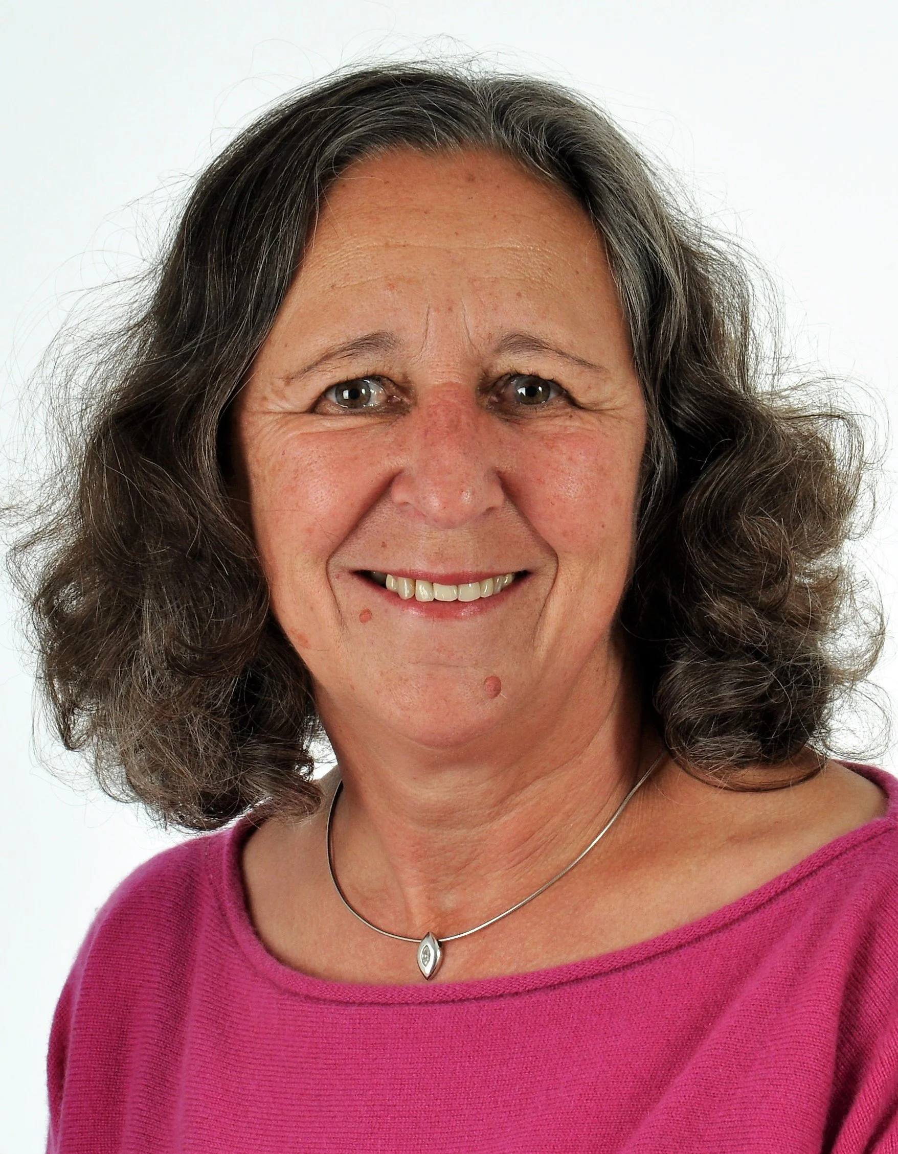 Nahaufnahme einer smiling Frau mit braunen, lockigen Haaren und einem pinken Oberteil, trägt einen silbernen Halsreif mit Anhänger, vor weißem Hintergrund.