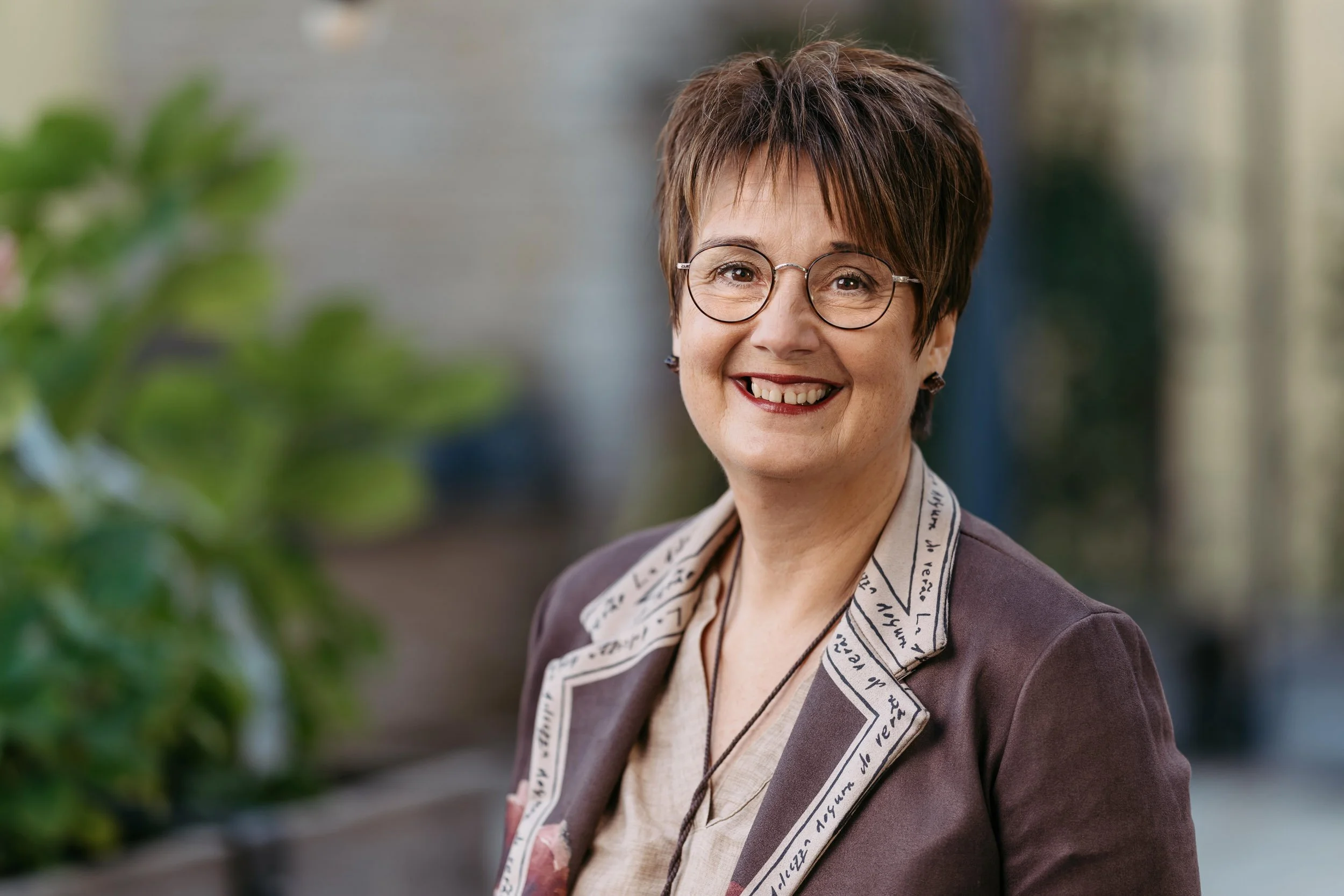 Eine lächelnde Frau mit kurzen braunen Haaren, Brille und einem braunen Blazer vor einem unscharfen Hintergrund.