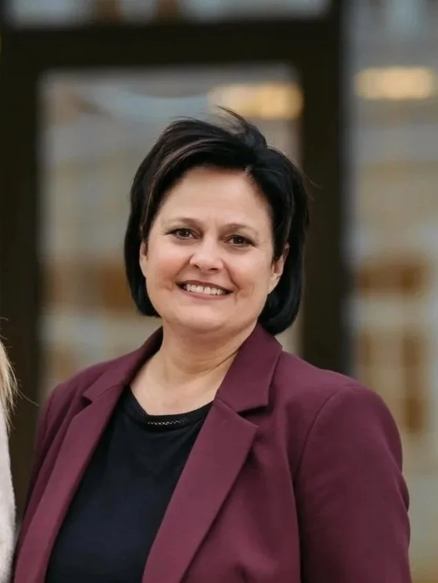 Eine Frau mit schwarzen Haaren, trägt eine dunkelrote Jacke und ein schwarzes Oberteil, lächelt vor einem Gebäude mit großen Fenstern.