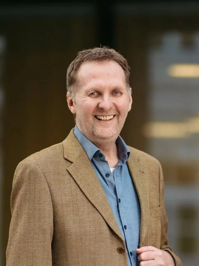 Ein lachender Mann in einem braunen Blazer und blauer Hemd, im Hintergrund unscharfe Bürolandschaft.