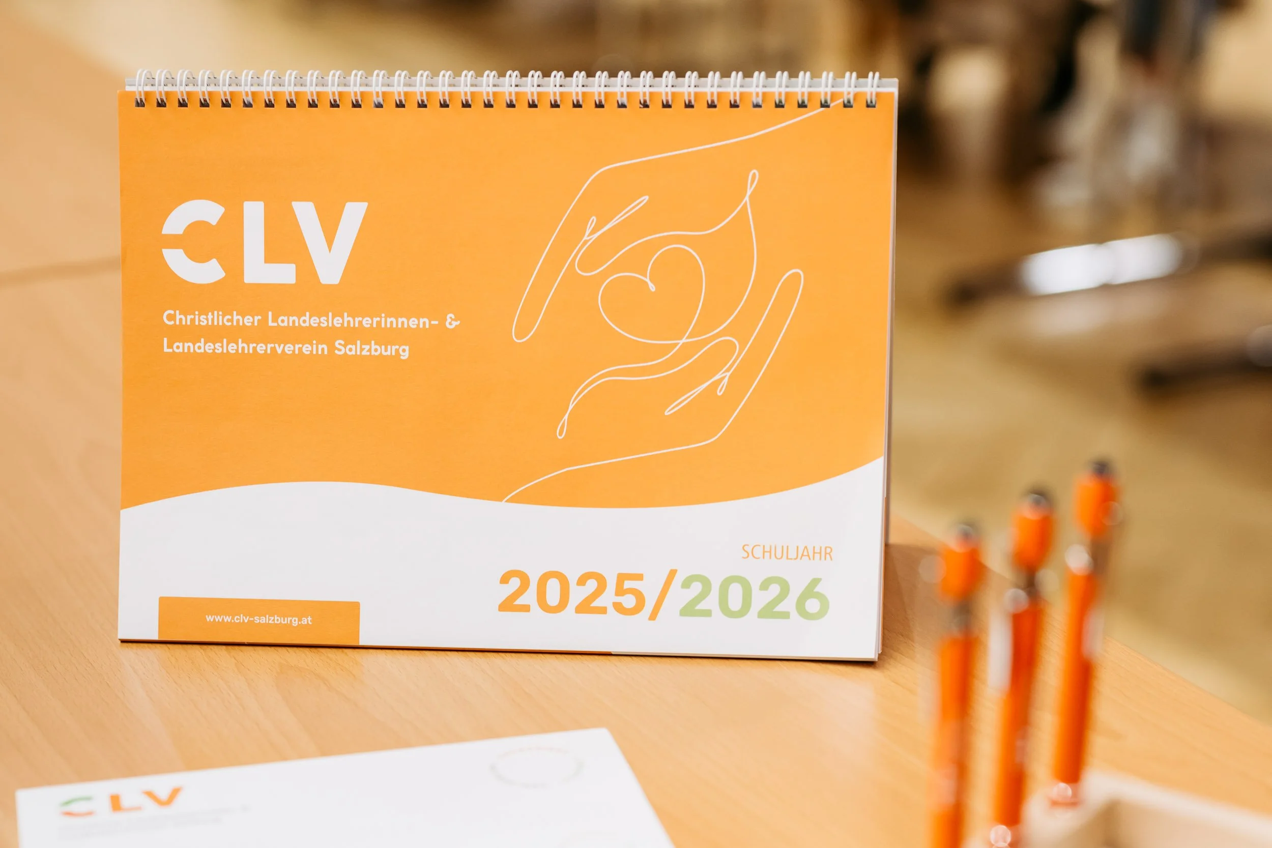 Schulkalender der Christlichen Landessenlehrerinnen- und Landessenlehrerverein Salzburg für das Schuljahr 2025/2026, in orange und weiß gestaltet, mit Illustrationen von zwei Händen, die ein Herz halten.