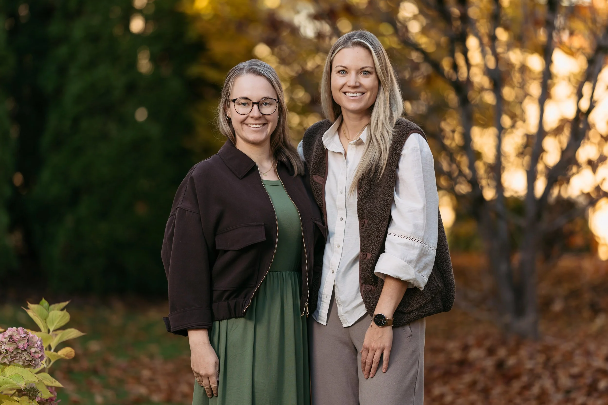 Zwei Frauen stehen im Freien im Herbst, lächeln und umarmen sich, umgeben von buntem Herbstlaub und Bäumen im Sonnenuntergang.