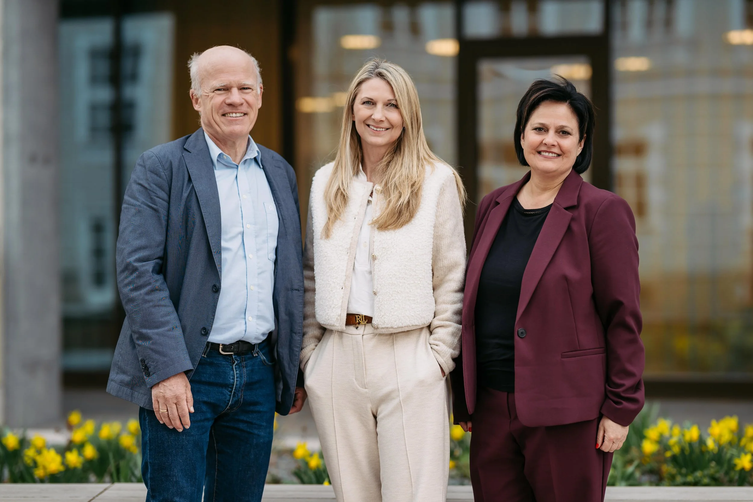 Drei Menschen, zwei Frauen und ein Mann, stehen nebeneinander vor einem modernen Gebäude und lächeln in die Kamera.
