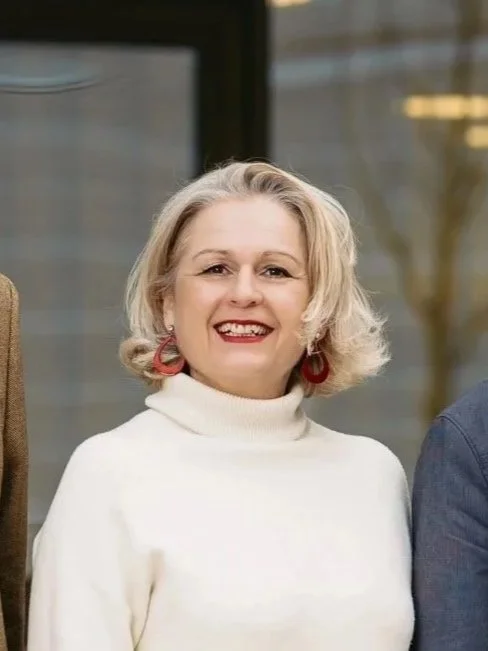 Lächelnde Frau mit kurzen, gewellten blonden Haaren, weißem Rollkragenpullover und roten Ohrringen, im Hintergrund ein Gebäude mit Fenstern.