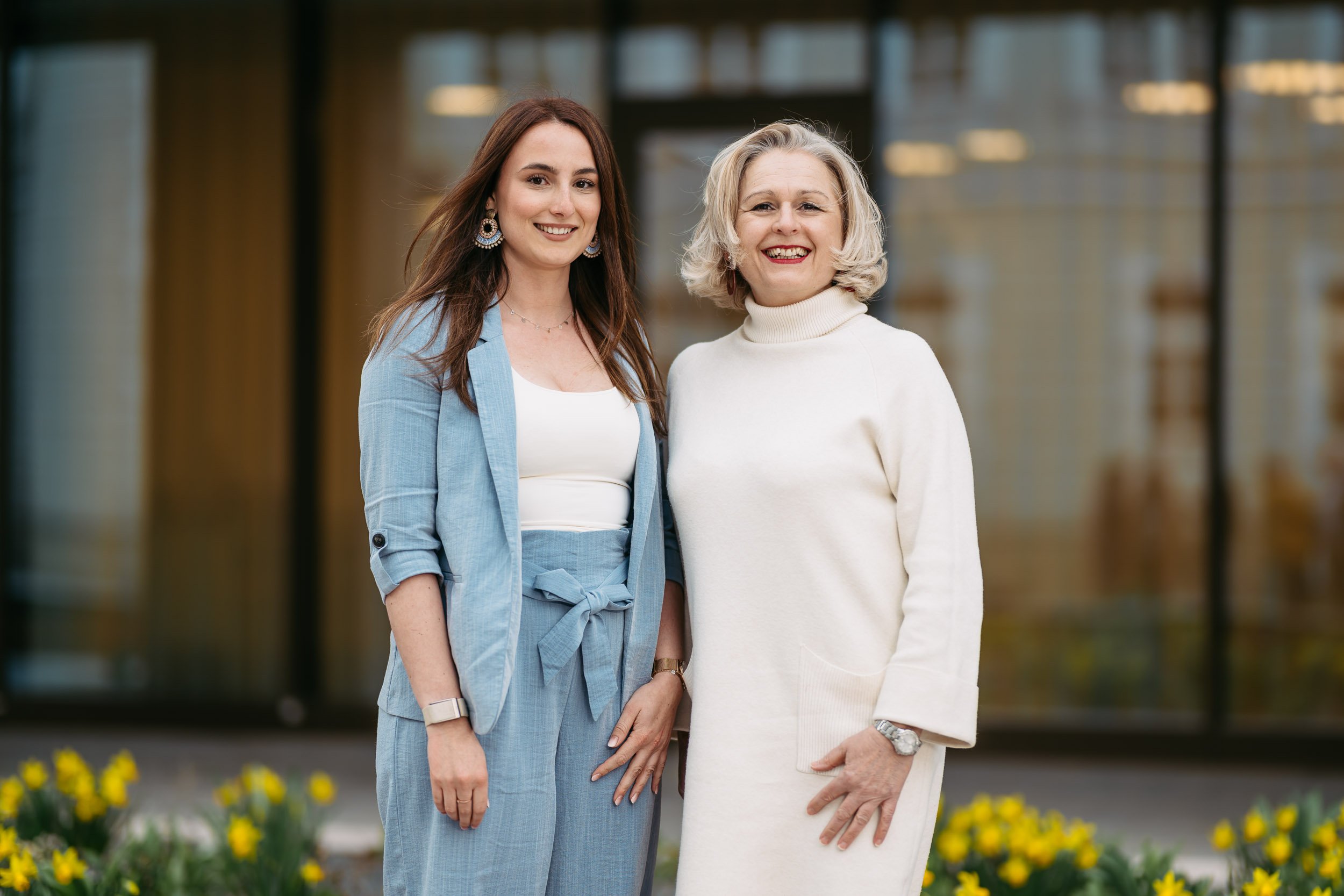 Zwei Frauen stehen nebeneinander draußen vor einem modernen Gebäude, lächeln in die Kamera, eine trägt ein hellblaues Business-Outfit, die andere ein weißes Kleid.