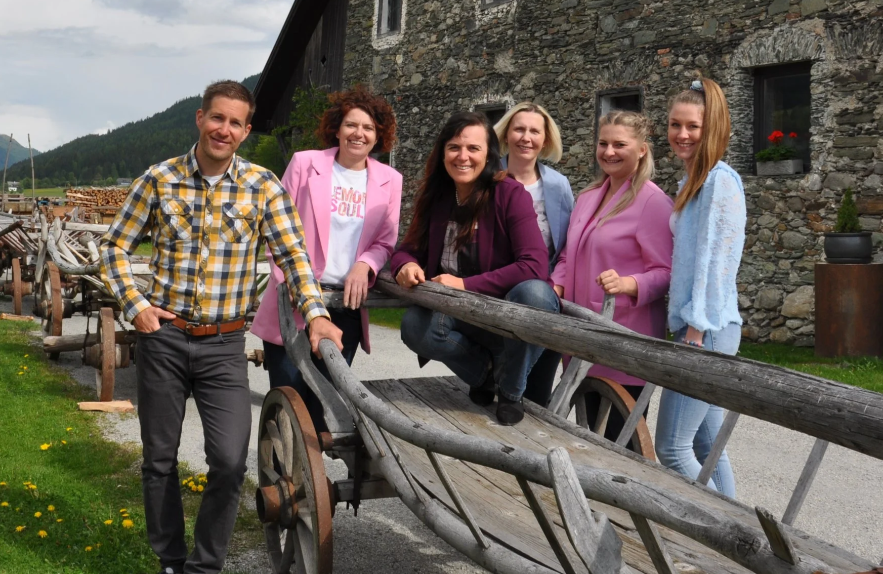 Sieben Menschen posieren draußen vor einem alten Bauernhaus mit Steingarten, die meisten tragen rosa oder blaue Jacken, und die Gruppe steht um eine alte Holzsperre. Im Hintergrund sind Berge und eine grüne Wiese sichtbar.