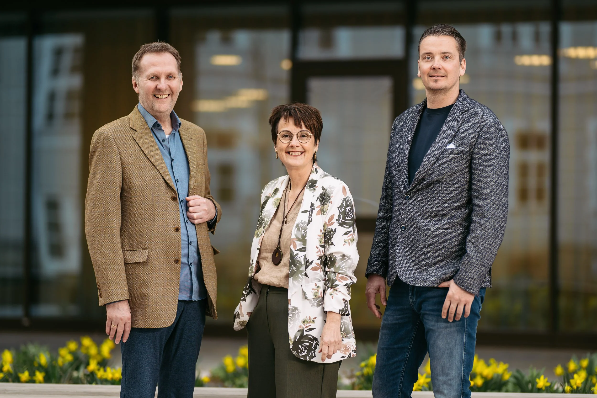 Drei Menschen stehen vor einem modernen Gebäude mit Glasfassade, alle lächelnd, im Freien mit Blumen im Vordergrund.