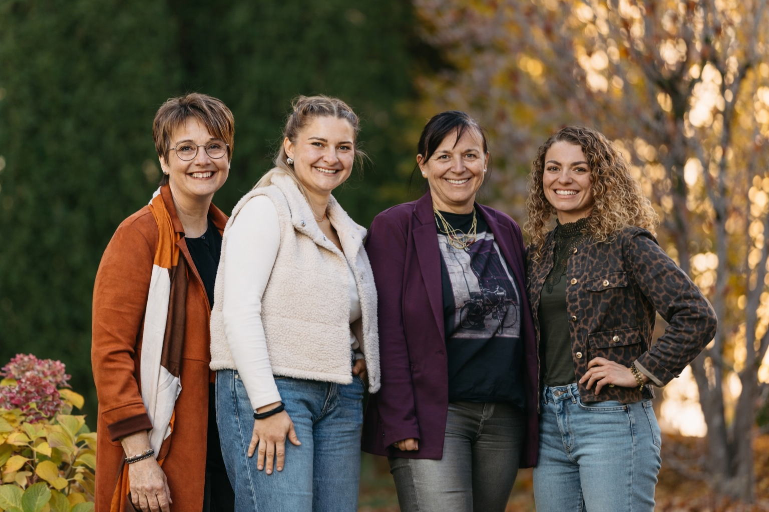 Vier Frauen stehen im Park im Herbst, lächelnd in die Kamera blickend.