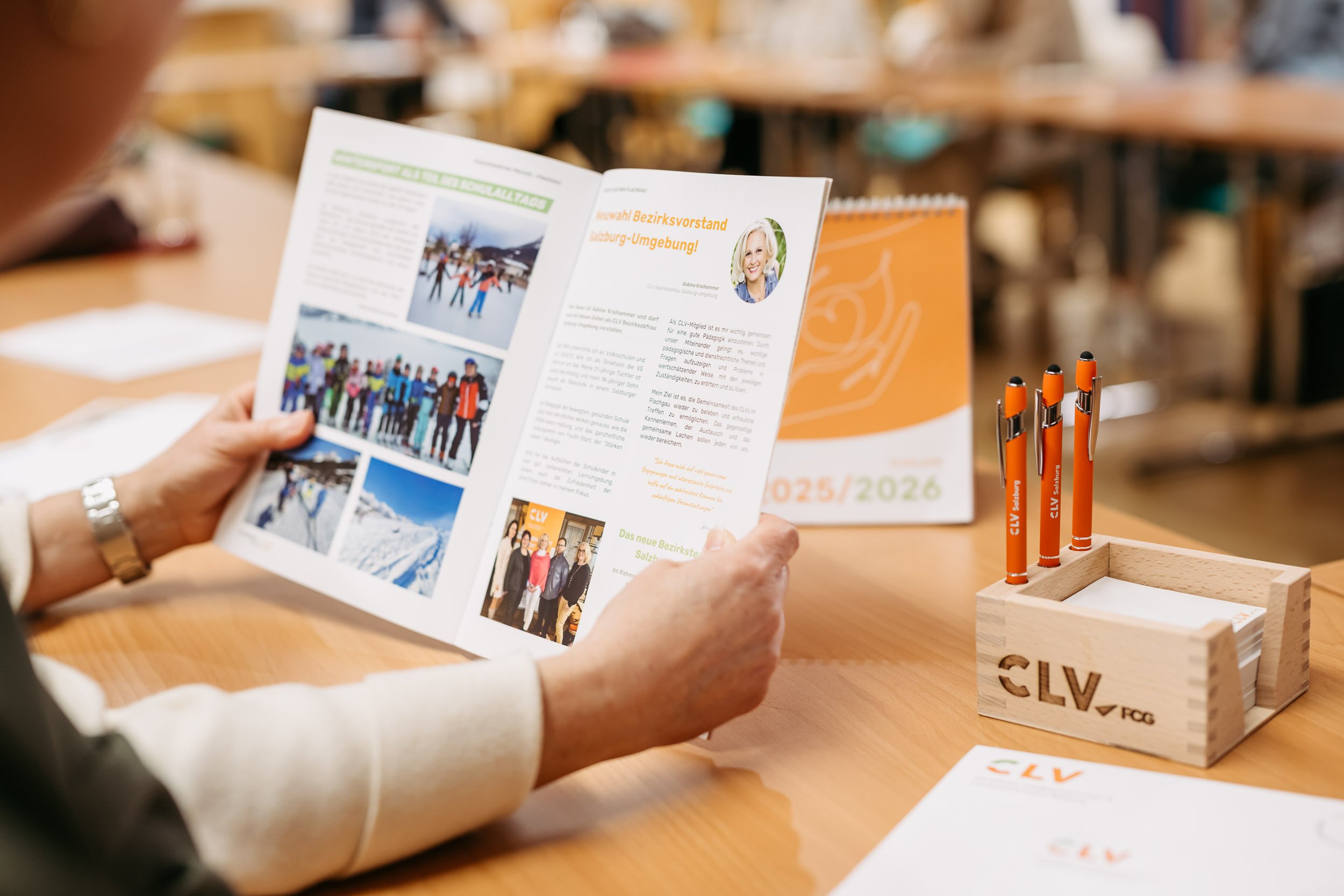 Eine Person liest eine Broschüre auf einem Holztisch in einer Geschäftsumgebung. Auf dem Tisch stehen orange Kugelschreiber in einem Holzständer und eine Karte mit dem Logo 'CLV'. Im Hintergrund sind weitere Tische und Stühle sichtbar.