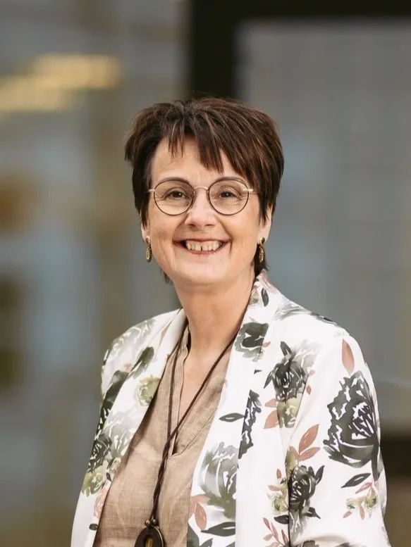 Eine lächelnde ältere Frau mit Brille, kurzen braunen Haaren und einem weißen Blazer mit Blumenmuster, im Hintergrund eine moderne Büroumgebung.