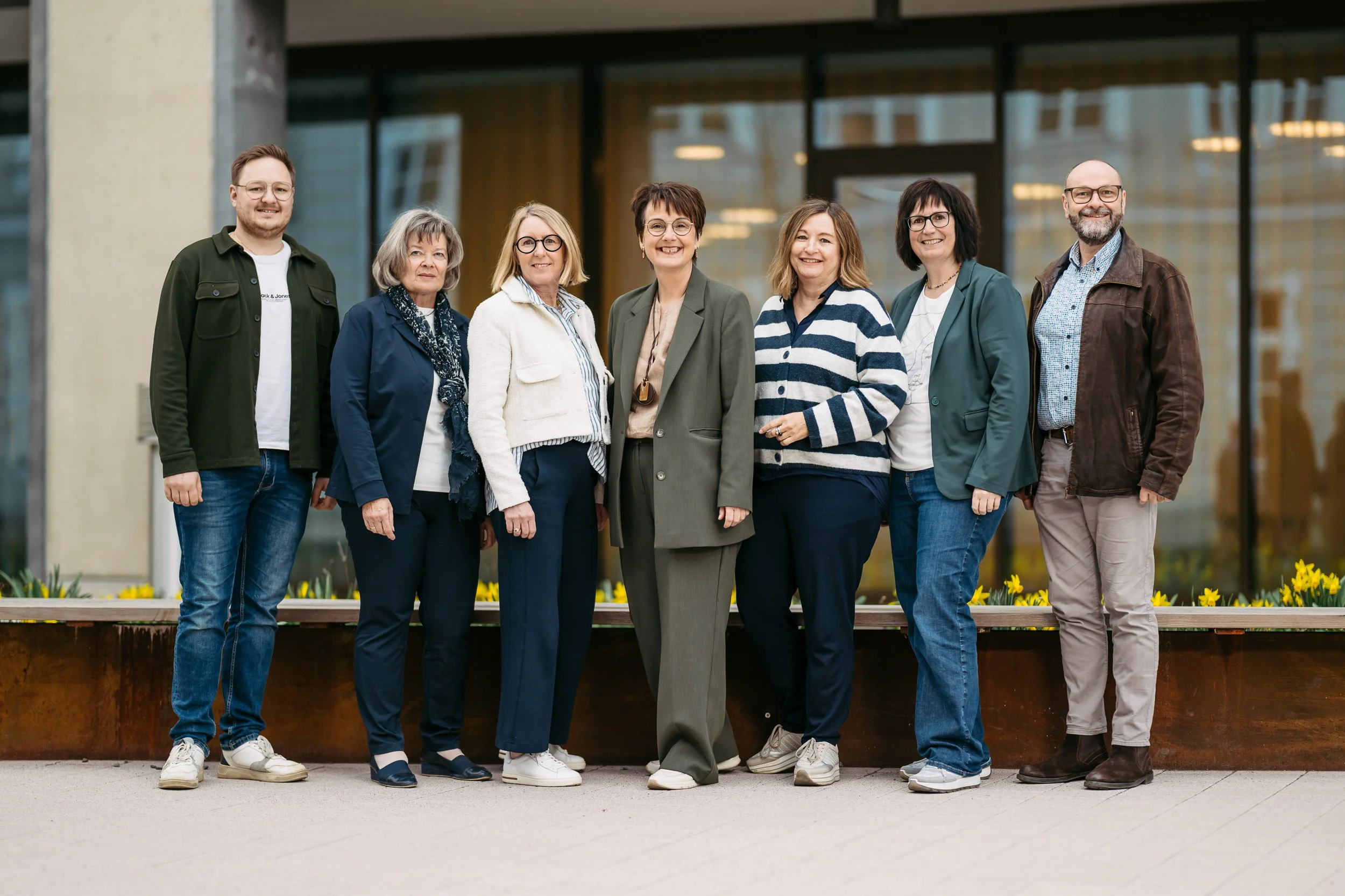 Gruppe von acht Menschen, die vor einem modernen Gebäude stehen, lachend und lächelnd, bei einer Gelegenheit wie einem Treffen oder einer Veranstaltung, bei sonnigem Wetter.