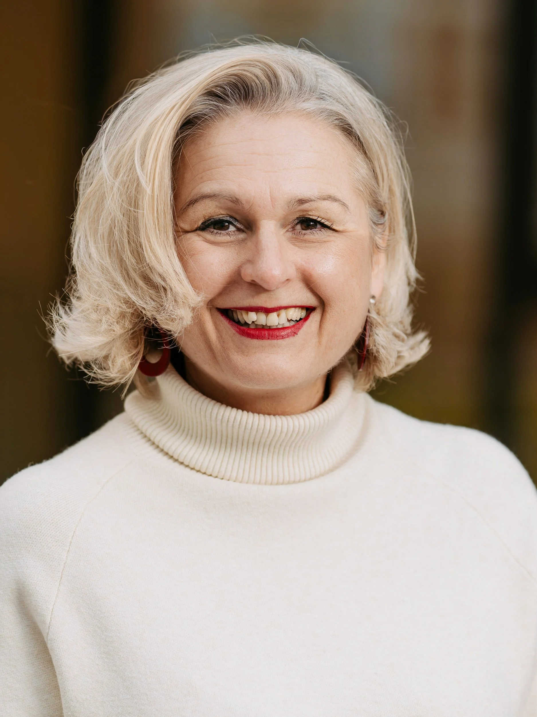 Nahaufnahme einer lächelnden älteren Frau mit blonden, lockigen Haaren, rote Lippen und weißen Zähnen, trägt einen weißen Rollkragenpullover.