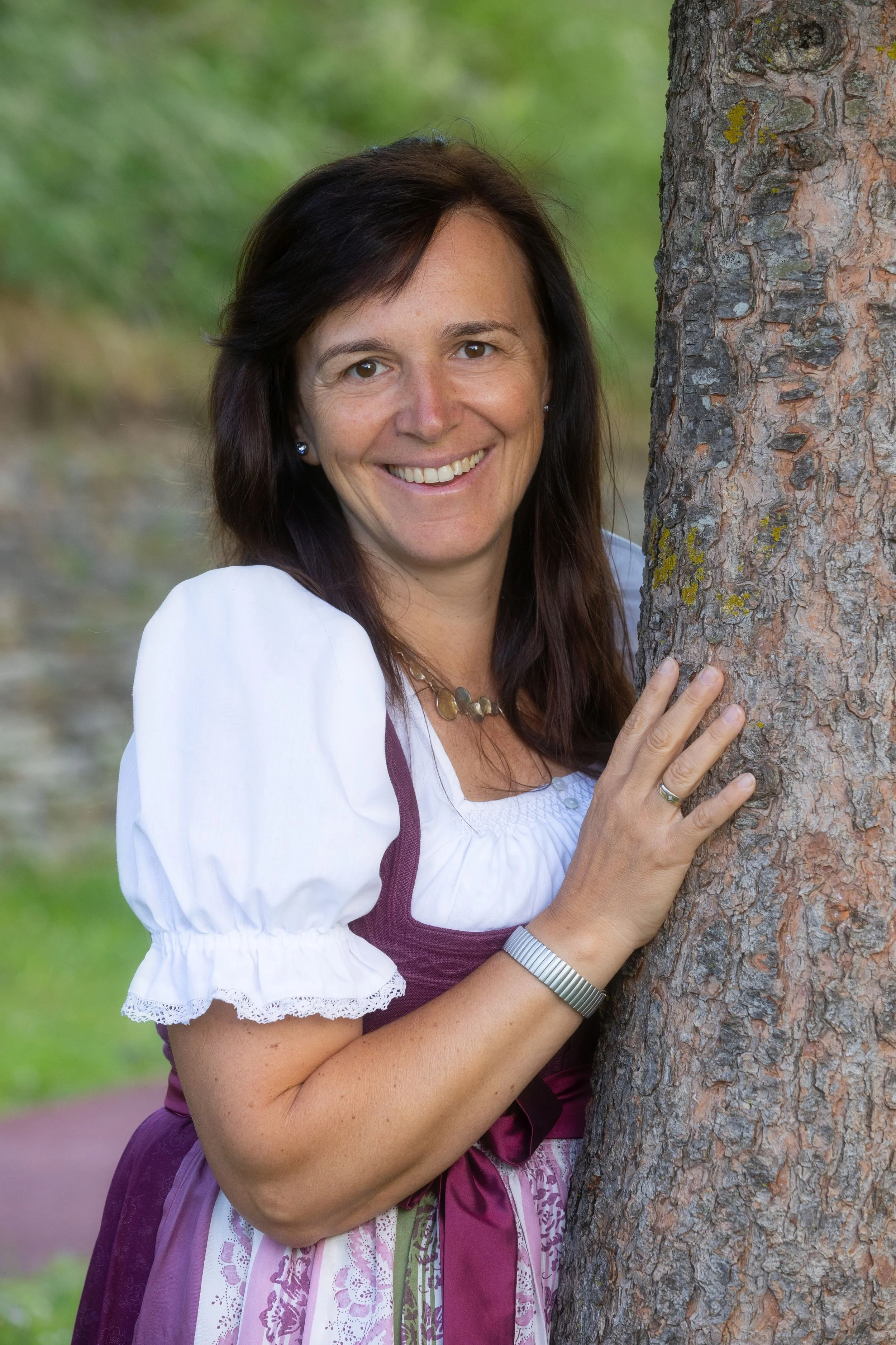 Eine lächelnde Frau in traditionellem deutschen dirndl trägt, die an einen Baum im Grünen lehnt.