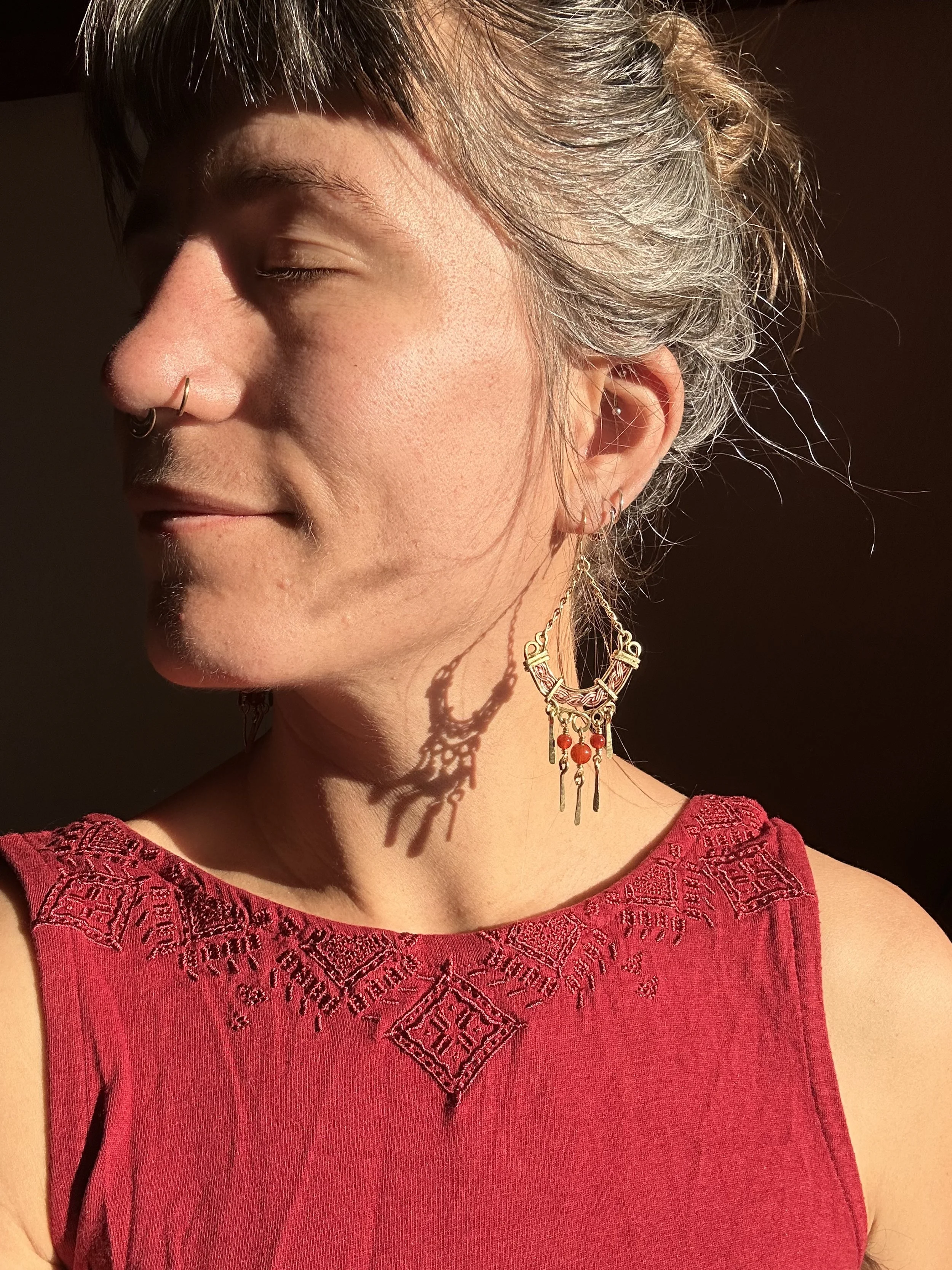 Femme portant une robe rouge avec broderie, portant des boucles d'oreilles dorées avec des perles rouges, avec ses cheveux gris attachés, la lumière du soleil crée une ombre de ses bijoux sur son cou.