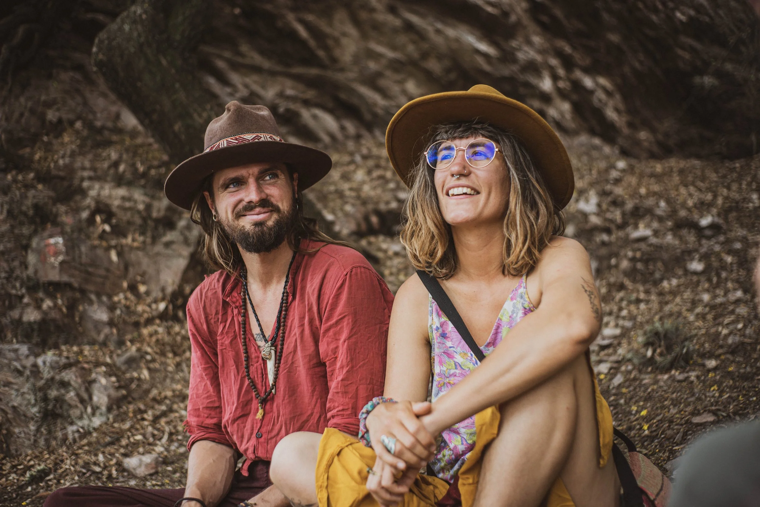 Deux personnes assises dans une forêt, portant des chapeaux, l'une avec des lunettes rondes, souriant et vêtue d'un débardeur coloré, l'autre avec une barbe et un chapeau, regardant vers elle, ambiance détendue.