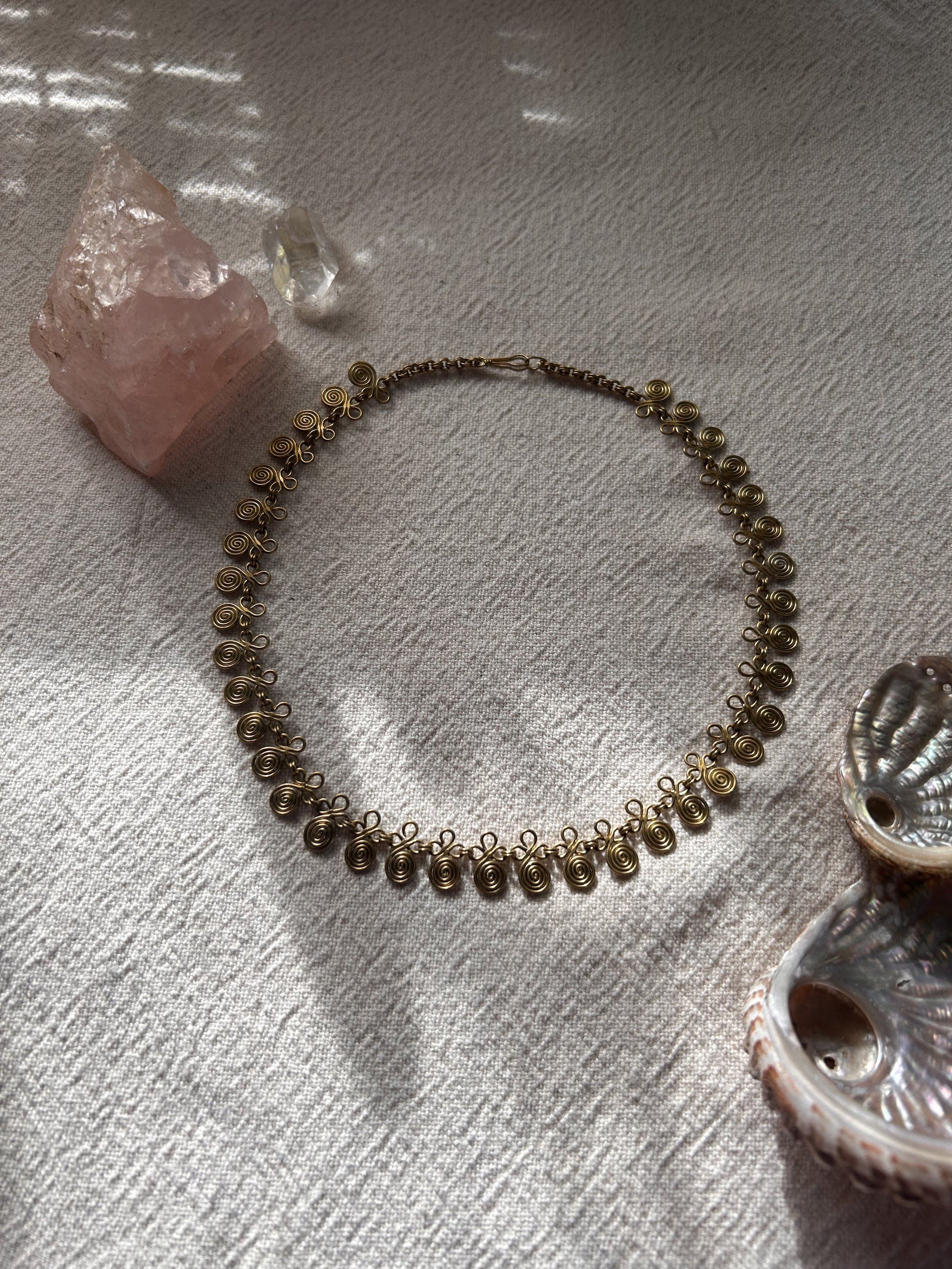 Collier en métal avec des spirales, posé sur une surface en tissu, entouré de cristaux et de coquillages.