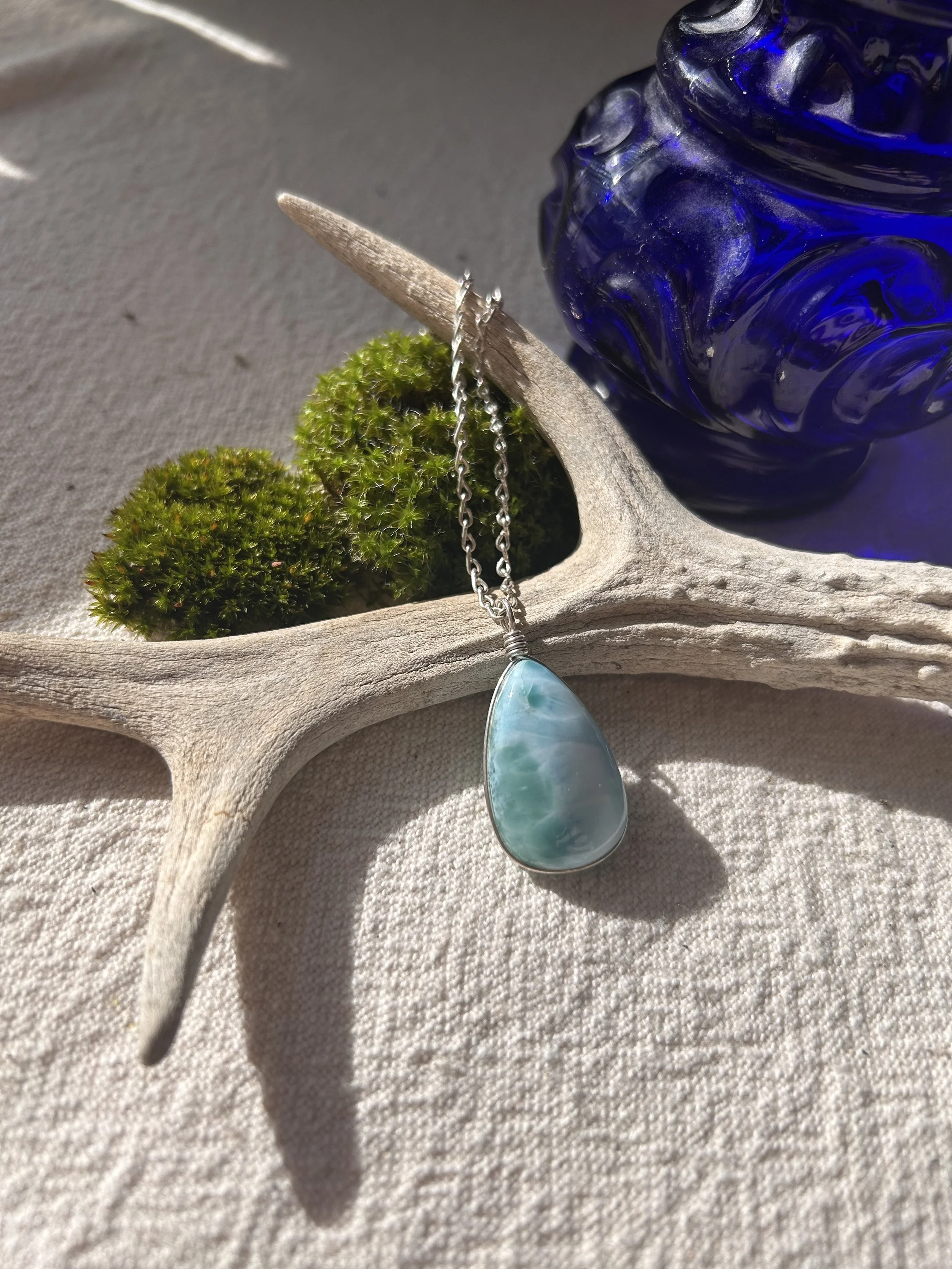 Collier avec pendentif en pierre bleue, posé sur un bois flotté, avec de la mousse verte et un vase en verre bleu en arrière-plan.
