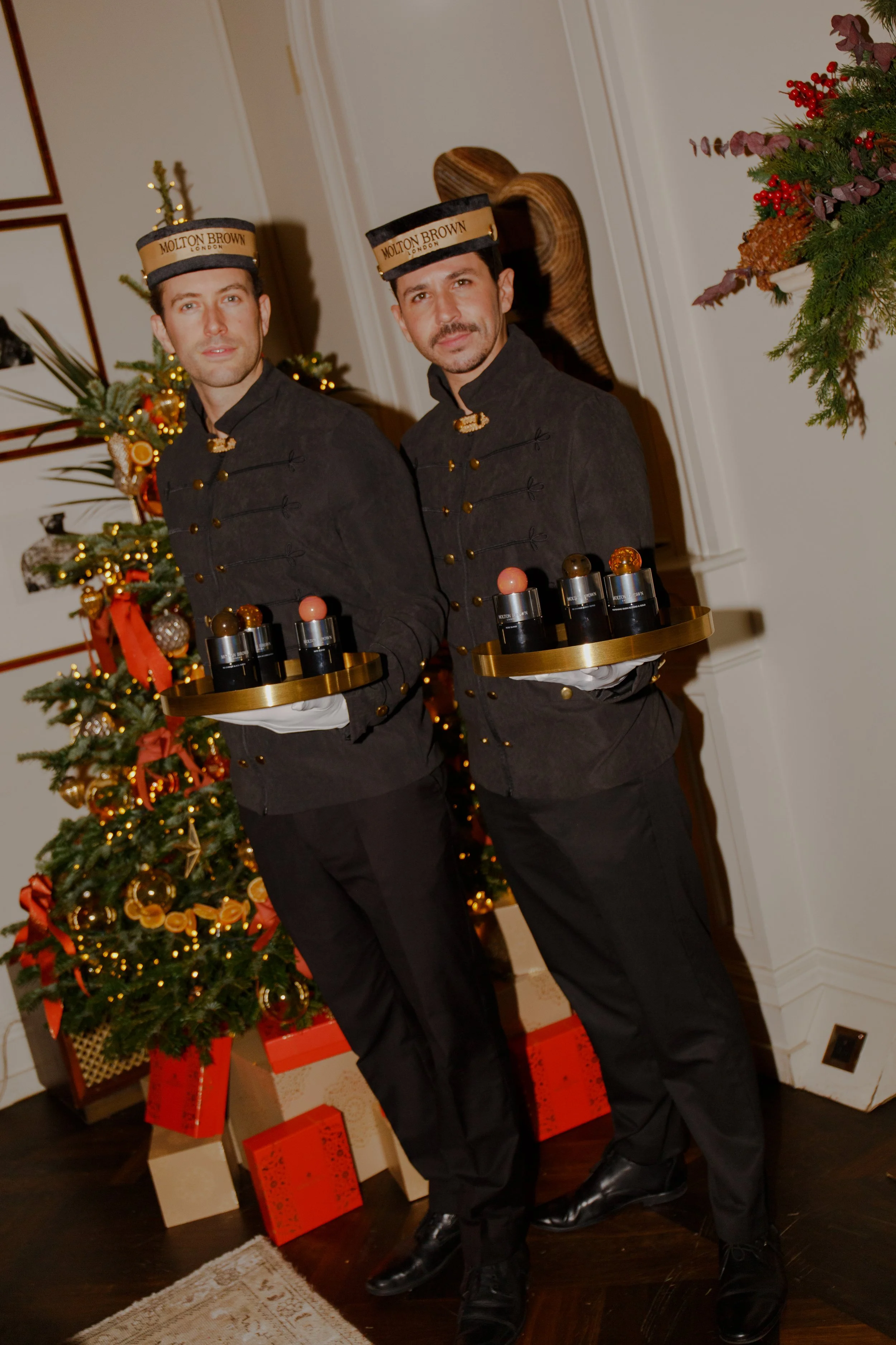 Two men dressed as bellhops are standing near a decorated Christmas tree, holding tray tables with Christmas ornaments. They are wearing black uniforms with brass buttons and hats labeled 'Molton Brown London.' The scene is festive with holiday decorations and wrapped gifts.
