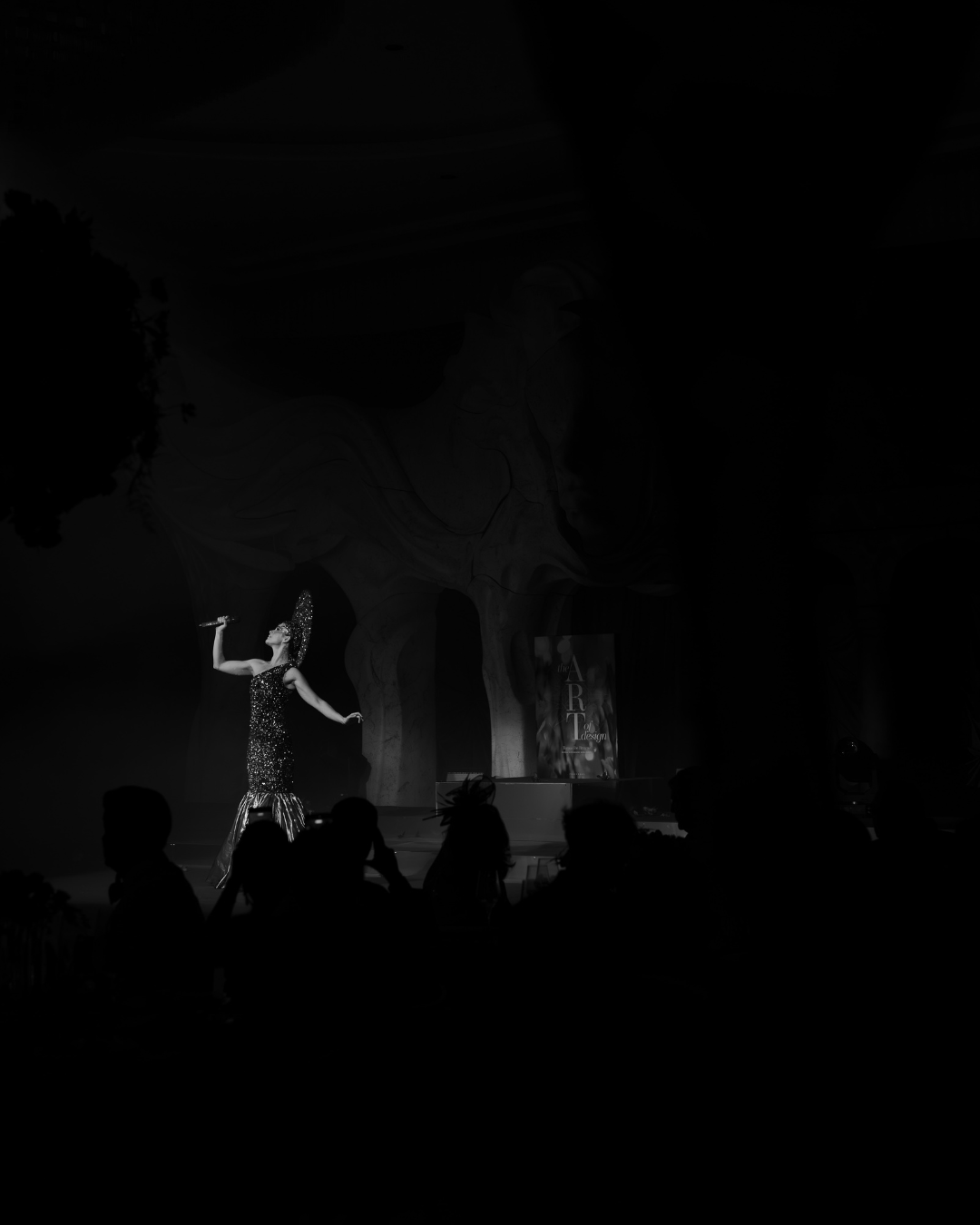 A performer dressed in a sparkling gown and headpiece singing on stage with audience silhouettes watching.