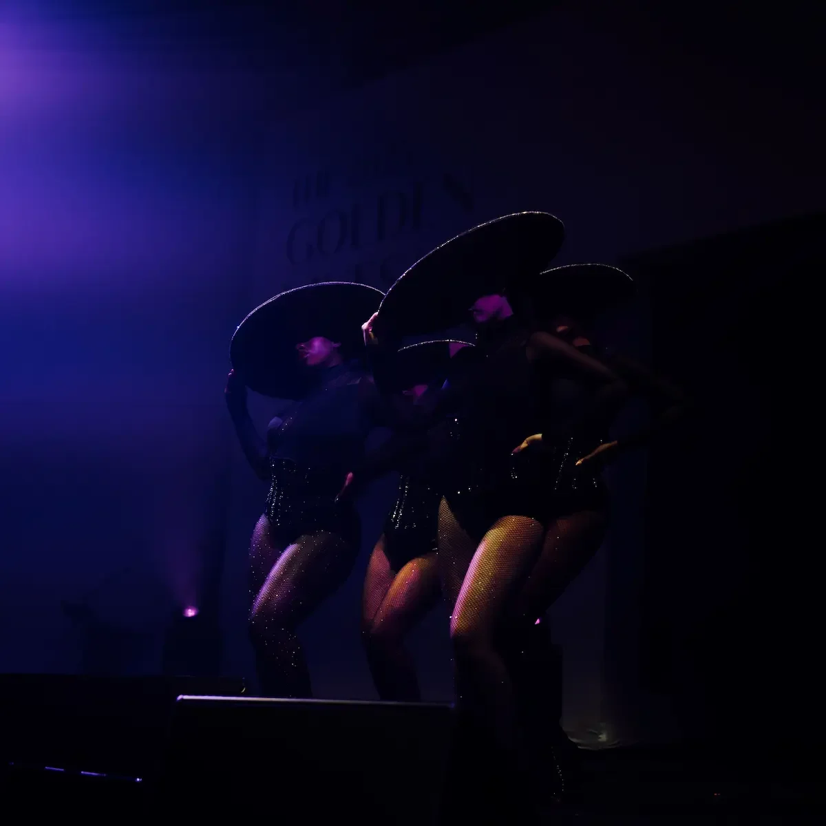 Three dancers in wide-brim hats performing under purple lighting