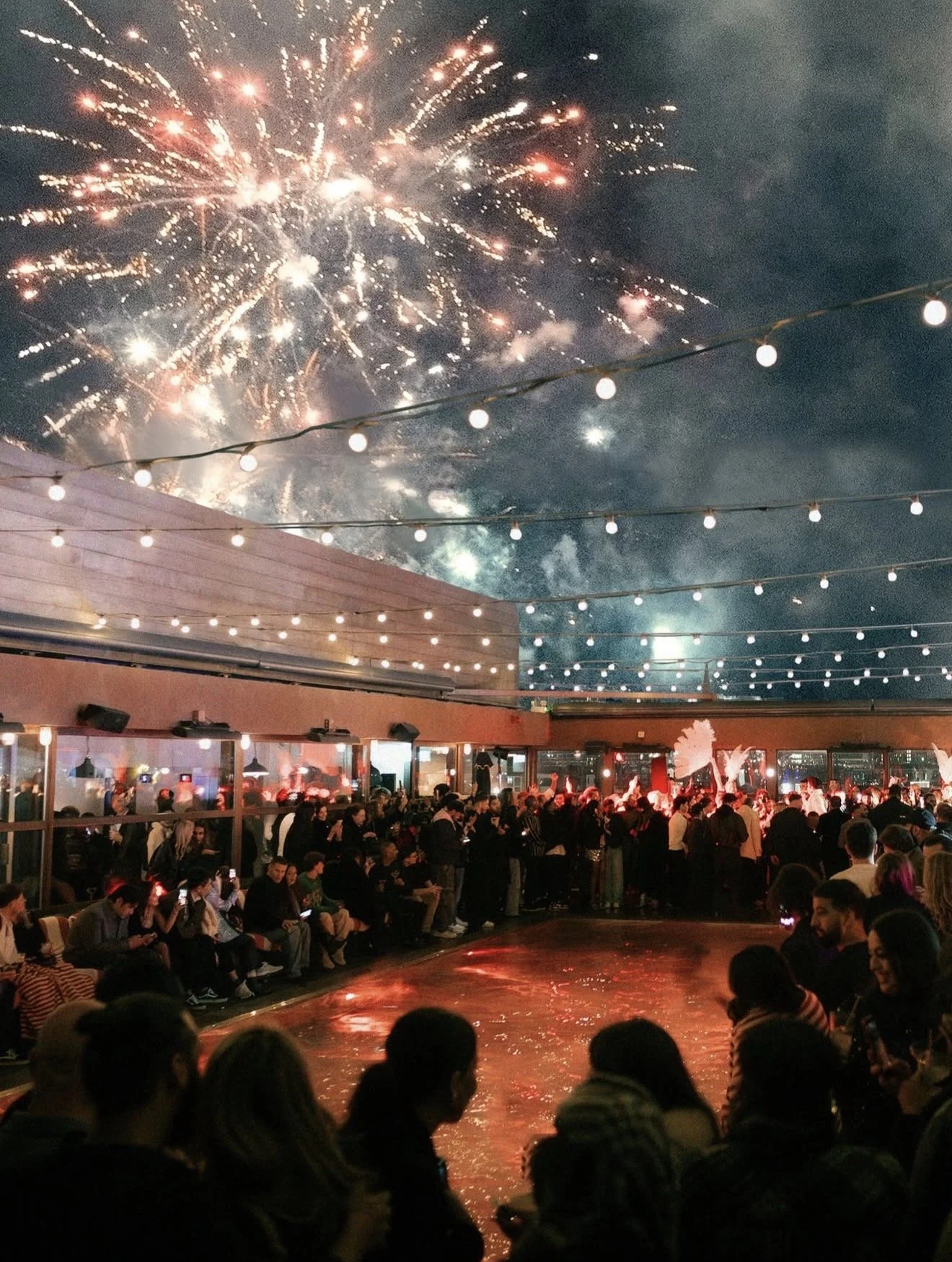 People gathered outdoors at night watching fireworks light up the sky. The scene is decorated with string lights and a water feature in the foreground, creating a festive atmosphere.
