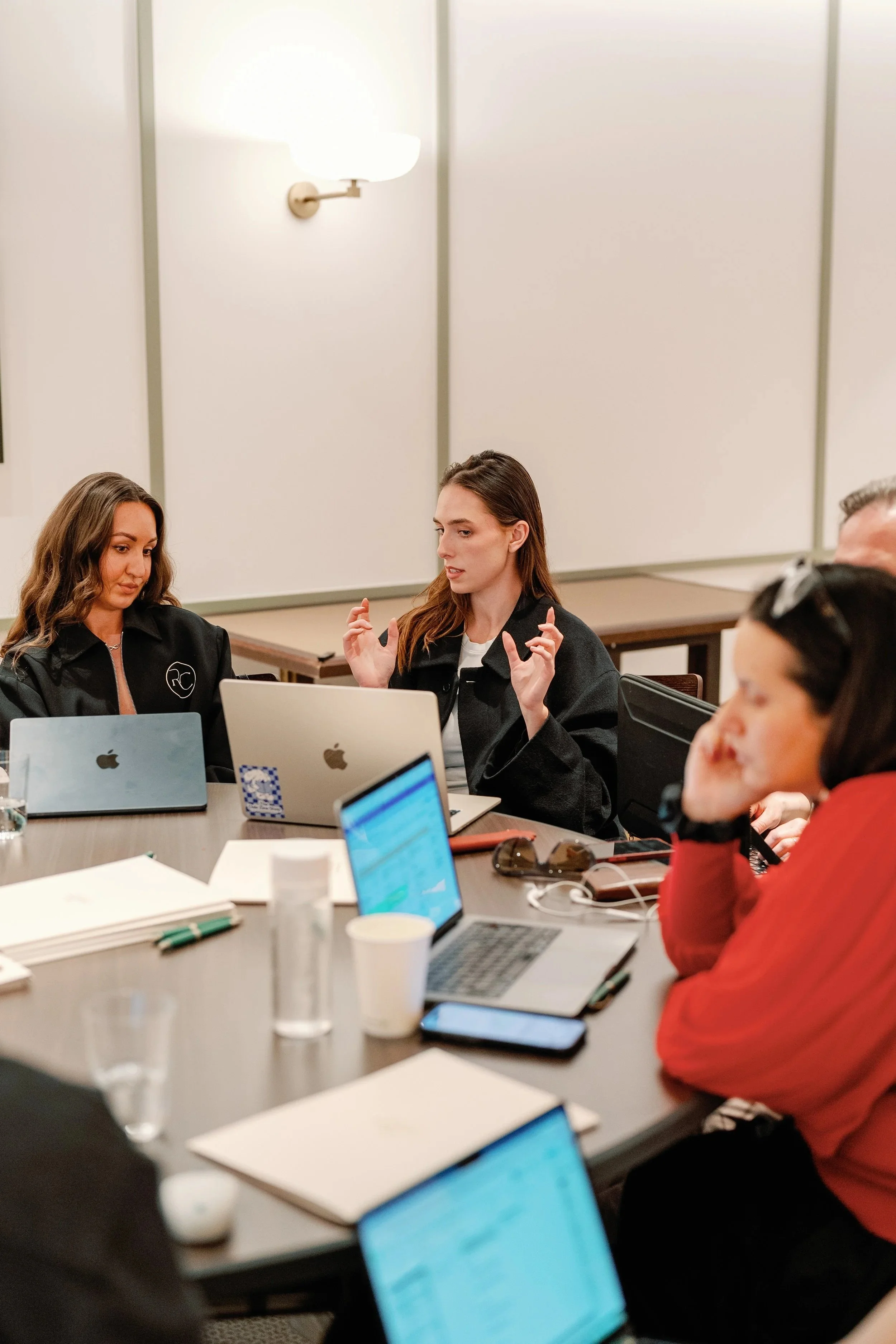 People sitting at a table with laptops, tablets, notebooks, and coffee cups in a meeting room, engaged in discussion.