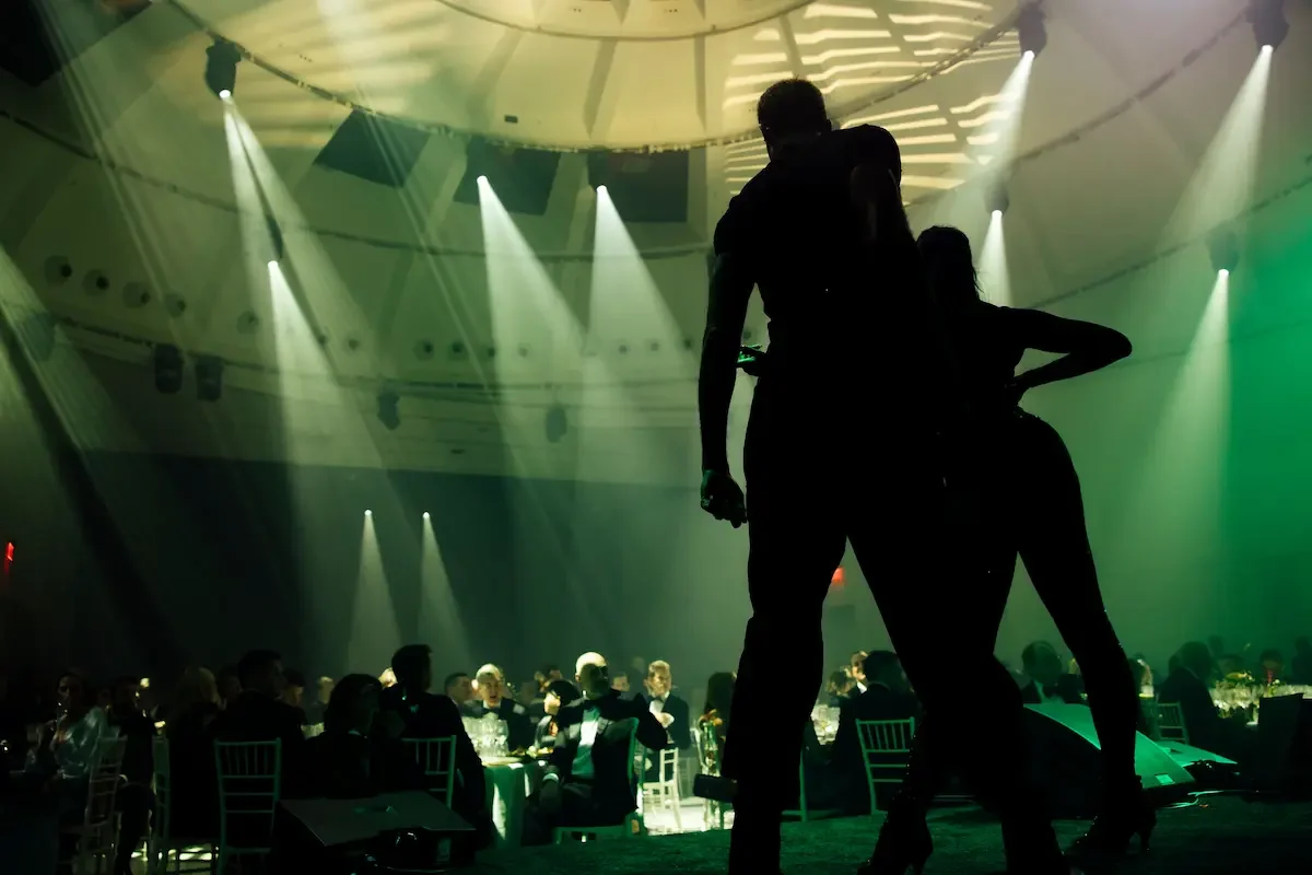 Dancing silhouettes on stage with green and yellow lighting and seated guests at a private event