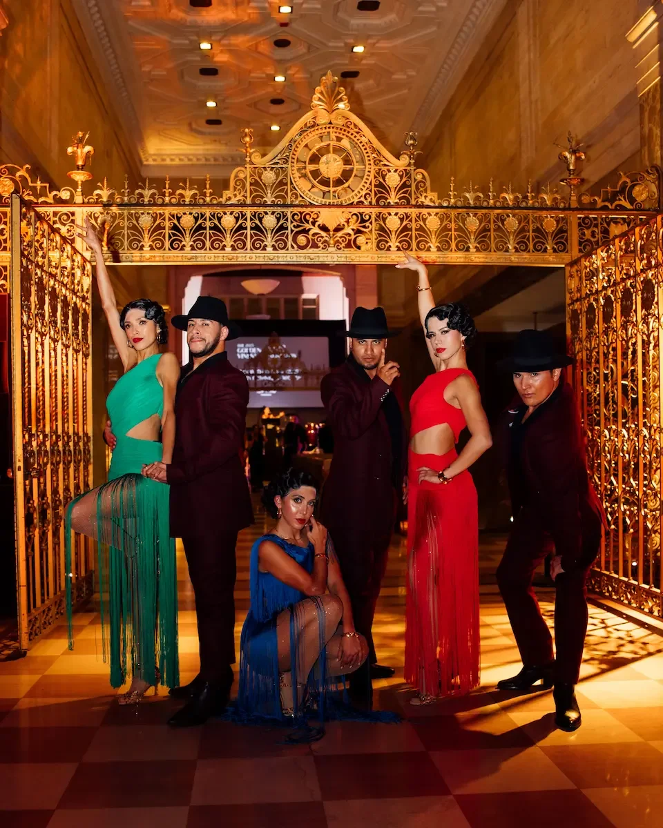 Five dancers in vibrant costumes inside gold-accented luxury foyer