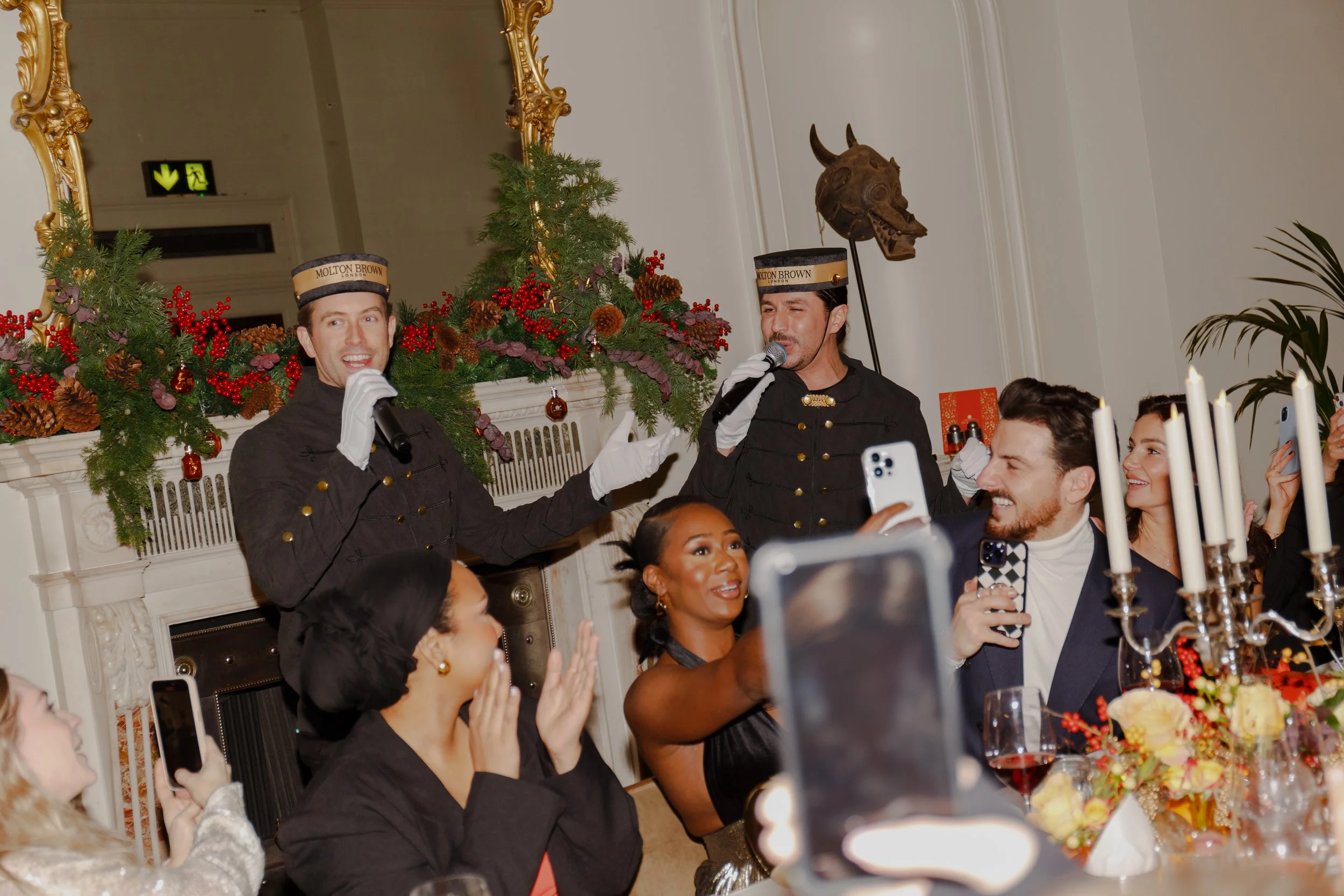 People dressed in vintage Clark Gable and Rhett Butler costumes singing at a Christmas party with festive decorations, candles, and guests taking photos.
