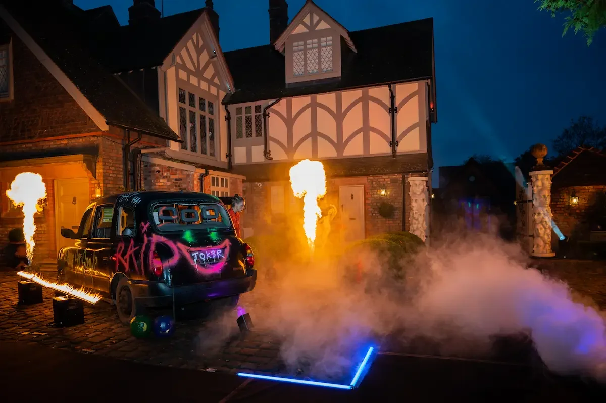 Graffiti-covered car with fire effects in front of a theatrical Tudor house