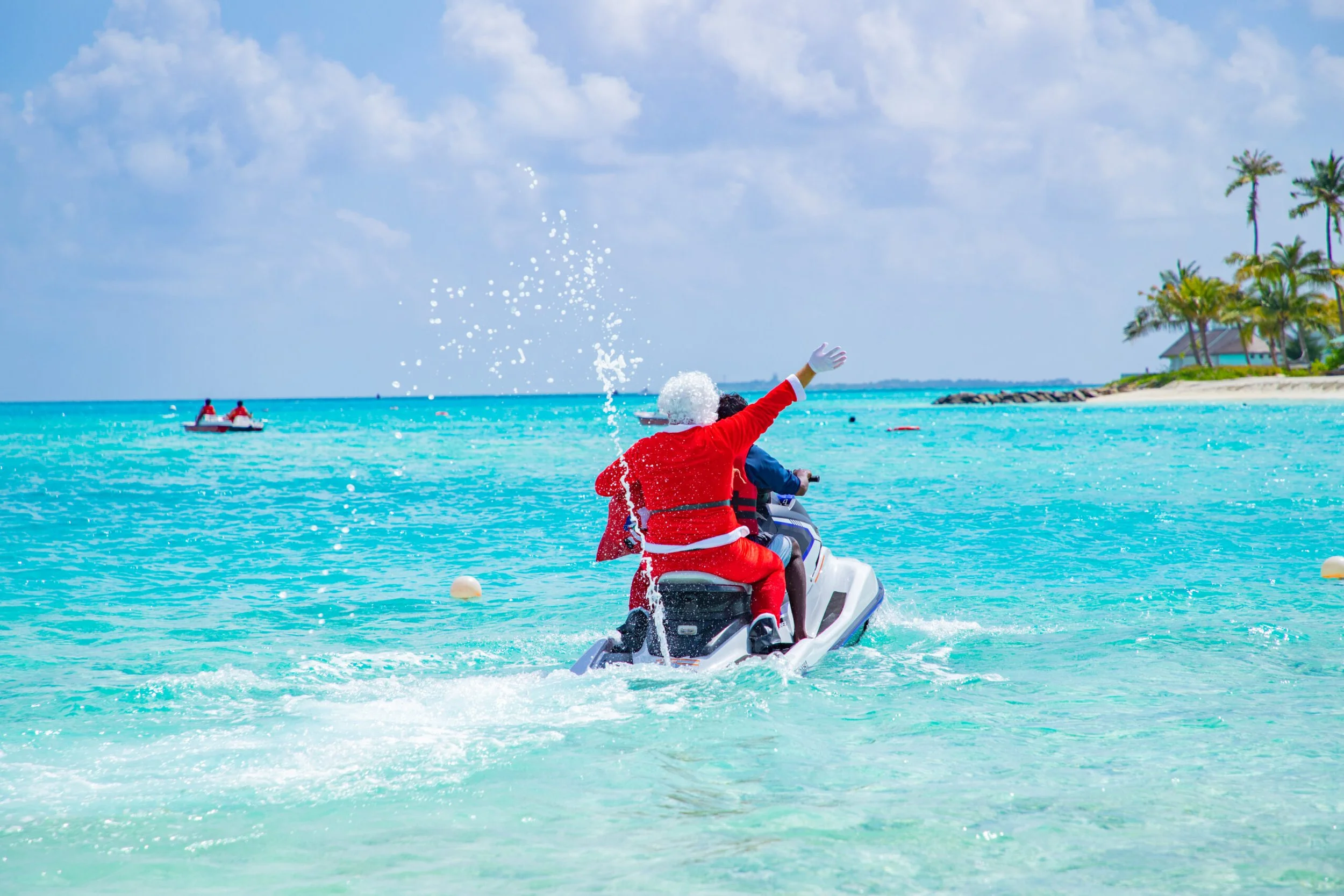 Person dressed as Santa Claus riding a jet ski on clear blue ocean waters with a festive splash, a boat in the distance, and a tropical island with palm trees in the background.