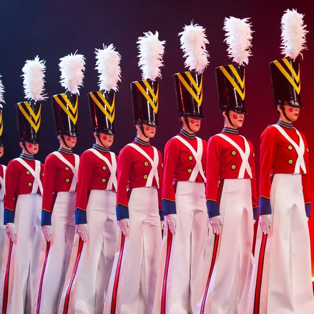 Line of dancers dressed as toy soldiers in red jackets with gold buttons, white pants with red stripe, tall black hats with yellow stripes and white plumes, and face paint with red cheeks.