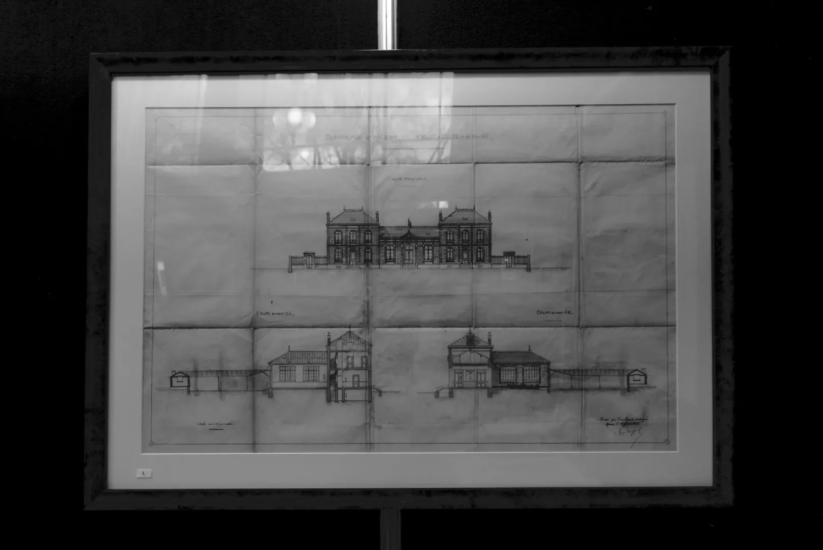 Black and white architectural blueprint of a large, historic building, displayed in a frame with reflections of lights and trees on the glass.
