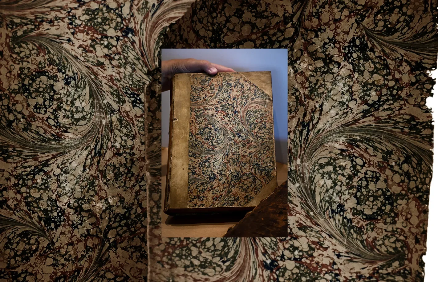 A person holding an old, marbled paper-covered book with a worn leather spine, with the book placed on a table with matching marbled paper.