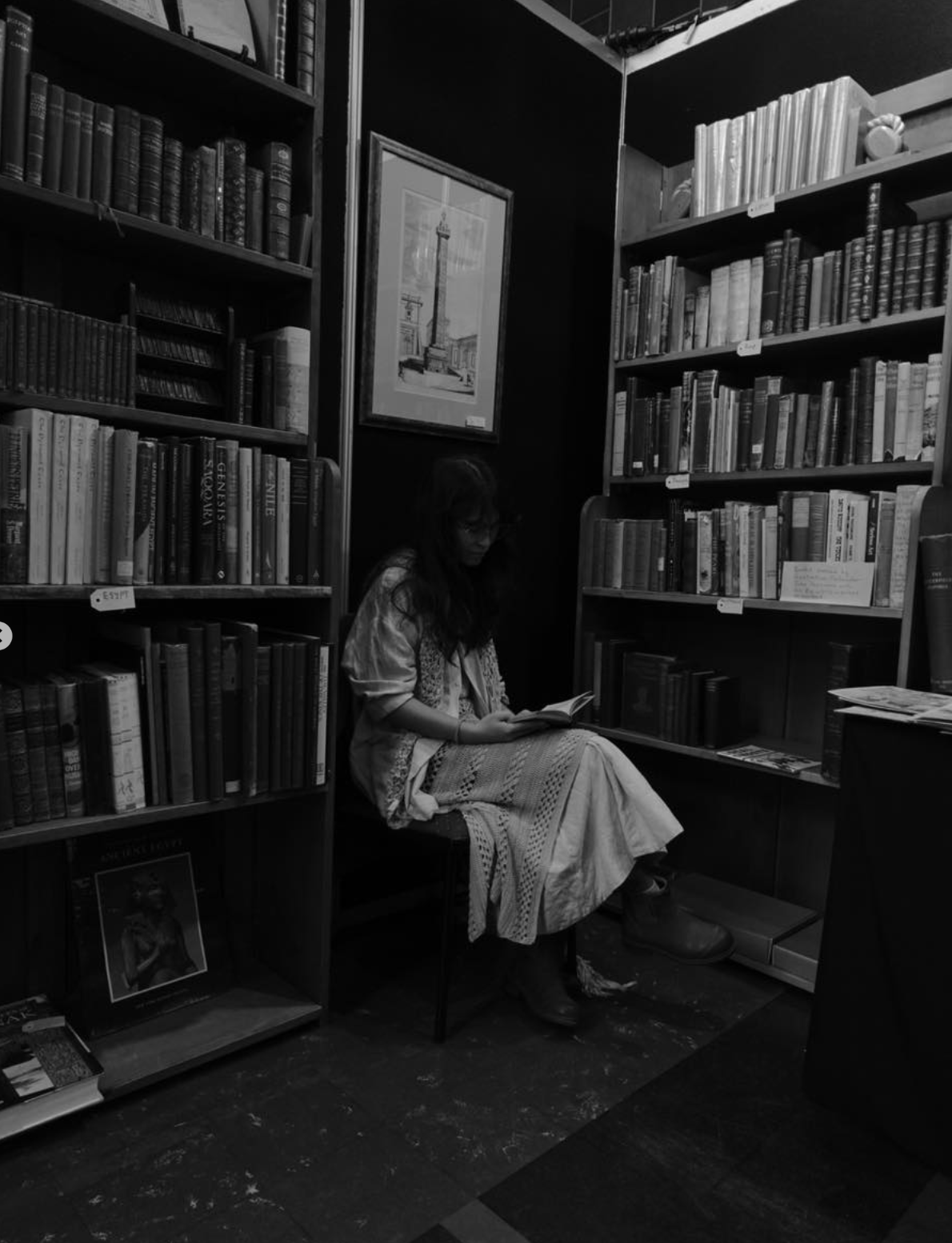 A woman sitting on a chair reading a book in a bookstore surrounded by tall bookshelves filled with books.