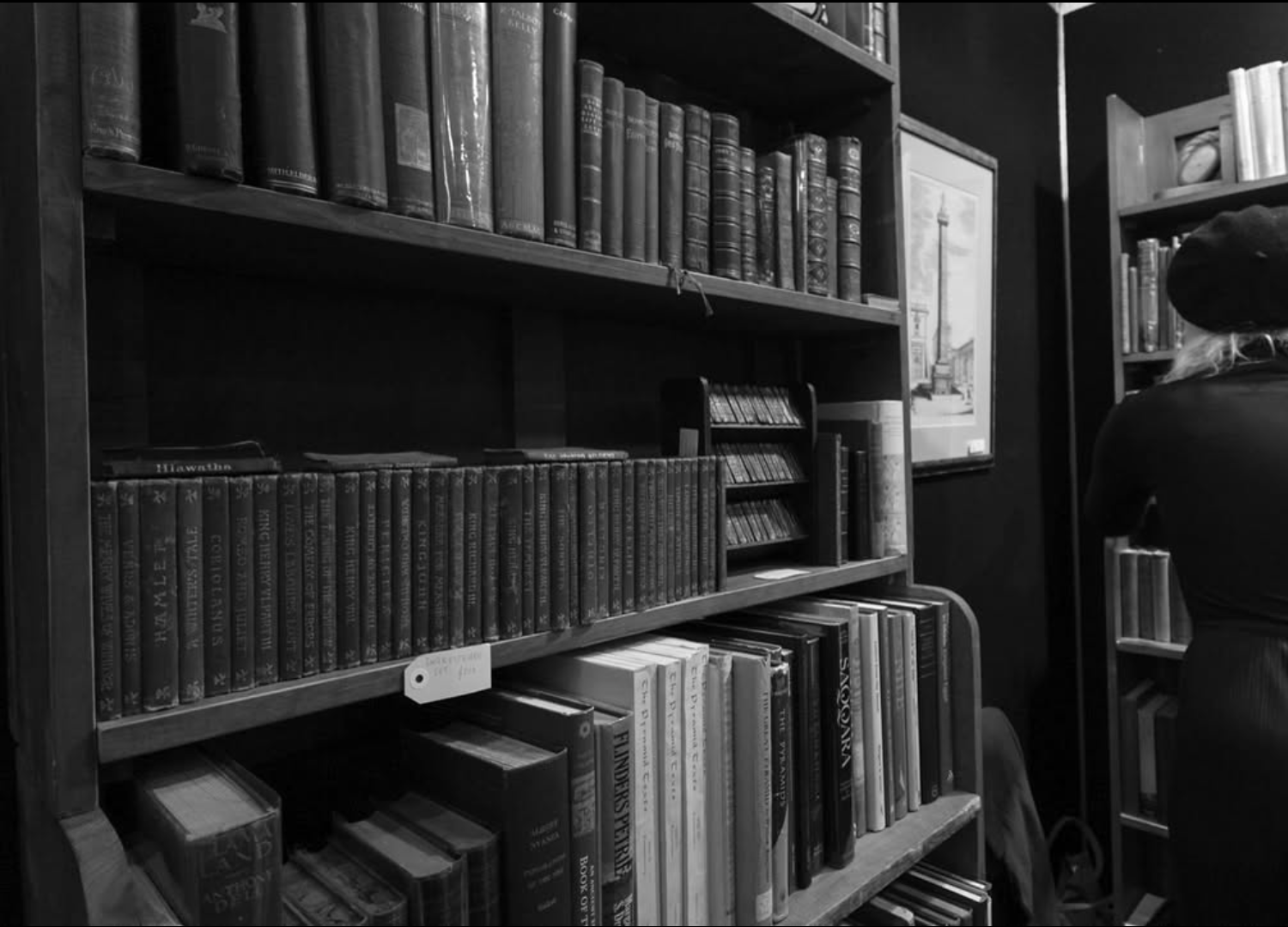 Black and white photo of bookshelf with books, a painting on the wall, and a person with long hair and a cap sitting on a chair.