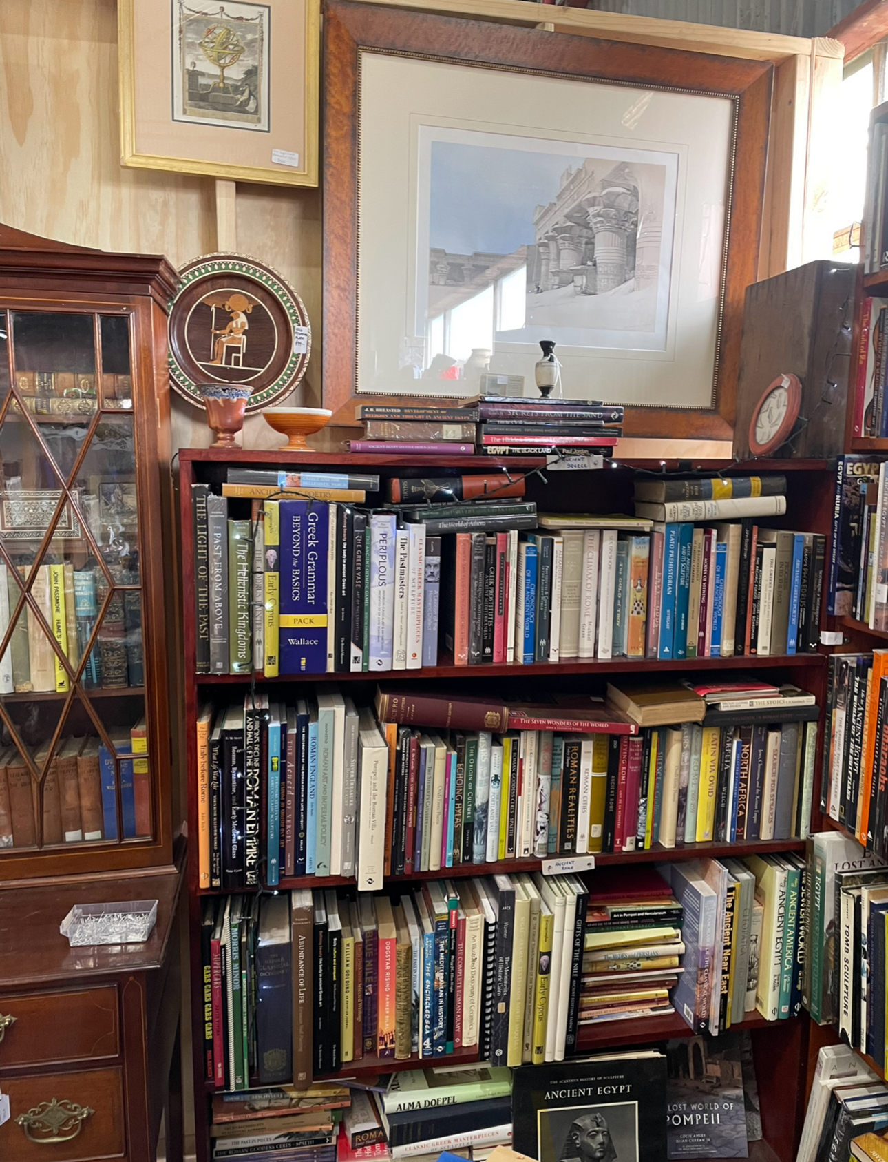 H20 Stall, at Waverley Antique Bazaar. Bookshelf's filled with a variety of books in a store, with framed artwork and decorative items on top and to the side.