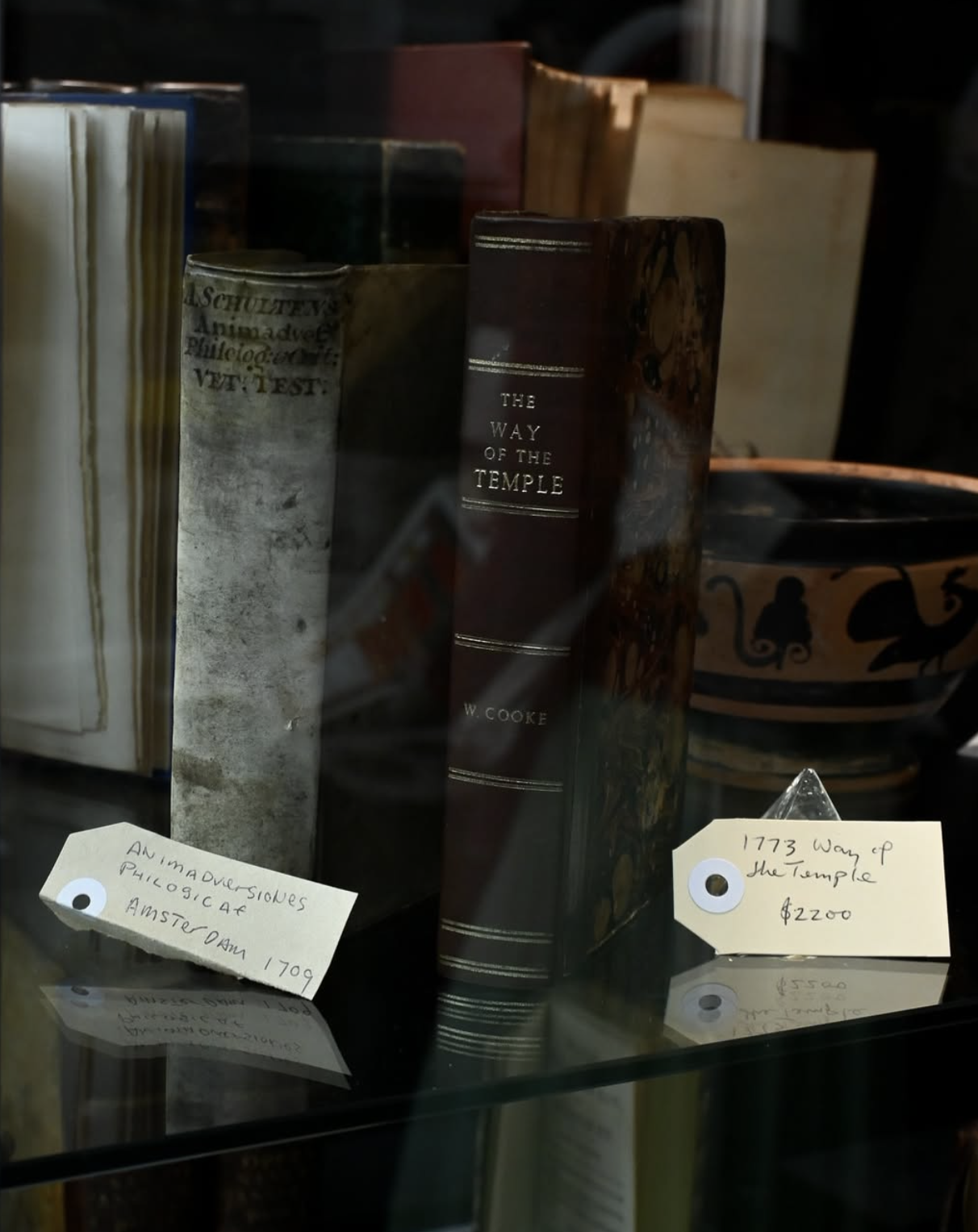 A display of antique books, including one titled 'The Way of the Temple' by W. Cooke, with handwritten tags indicating the books are from Amsterdam, 1709, and priced at $22.00.