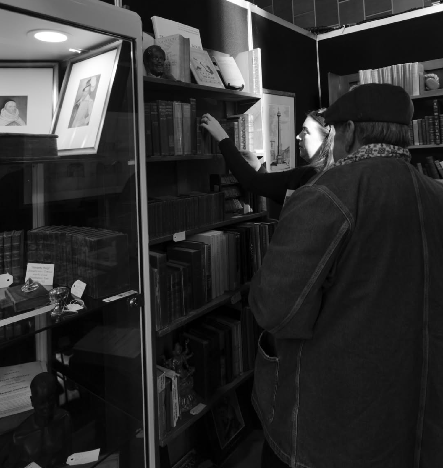 Two people browsing books in a bookstore or library. The woman is reaching for a book on a shelf, and the man is standing nearby, facing the shelves.