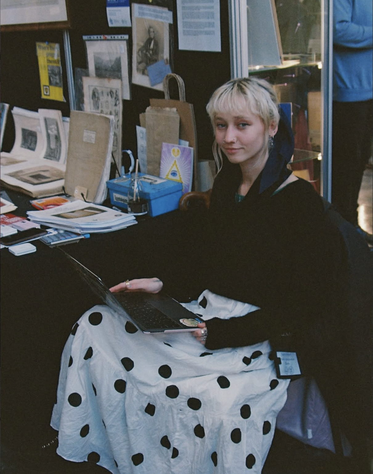 A woman with short blonde hair, sitting at a table with a black tablecloth, working on a laptop. The table has books, papers, and various items. Behind her are display boards with pictures and posters, and a brown paper bag. She is wearing a black top with a white skirt with black polka dots.