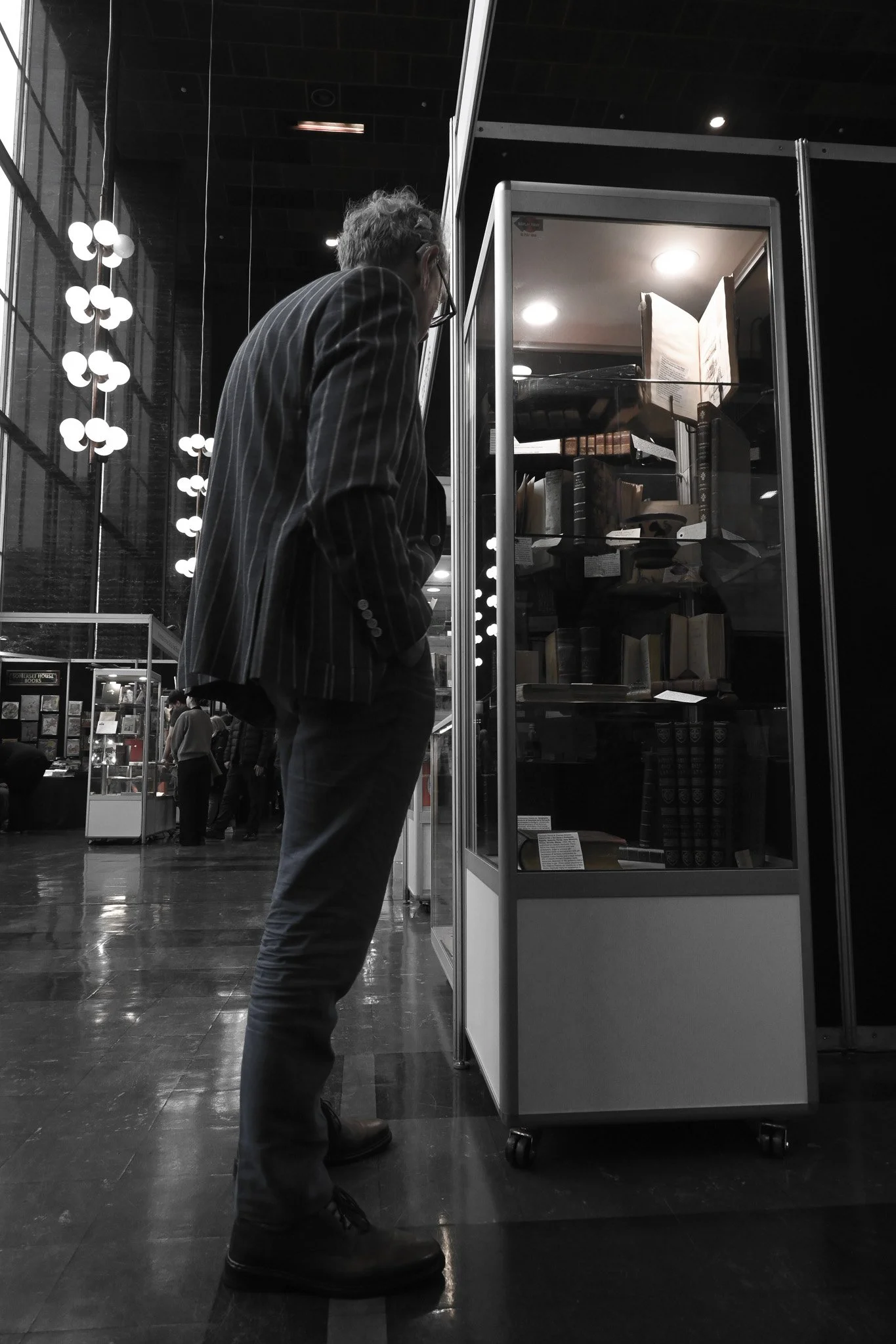 A man with glasses and a striped blazer looking at a glass display case with books inside at an indoor library or bookstore.