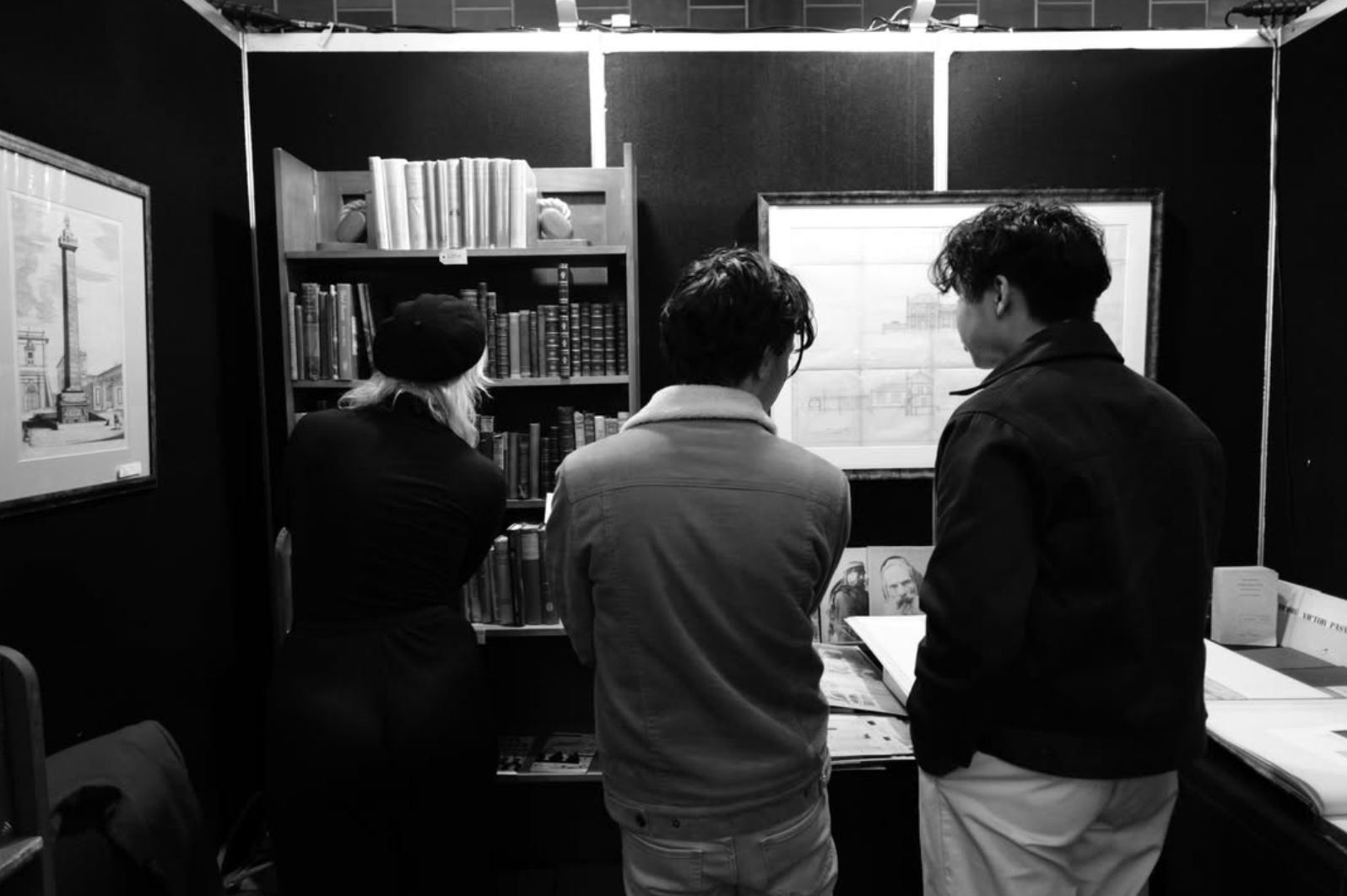 Three people looking at a book or artwork in a room with bookshelves and framed pictures on the wall.