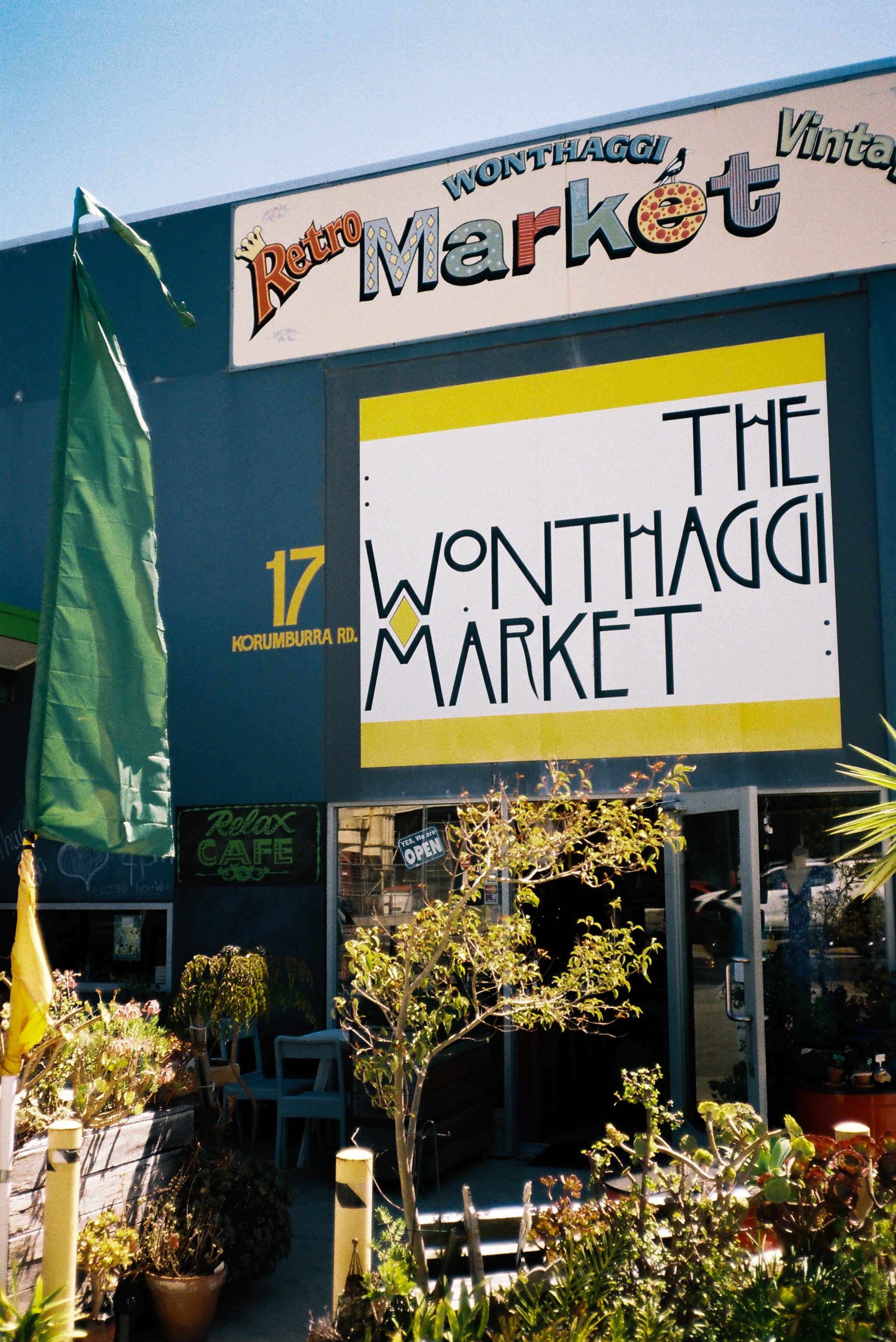 Exterior of Wonthaggi Market with sign reading 'The Wonthaggi Market' . There are plants and seating outside the entrance, with yellow and green flags and a blue building under a clear sky.