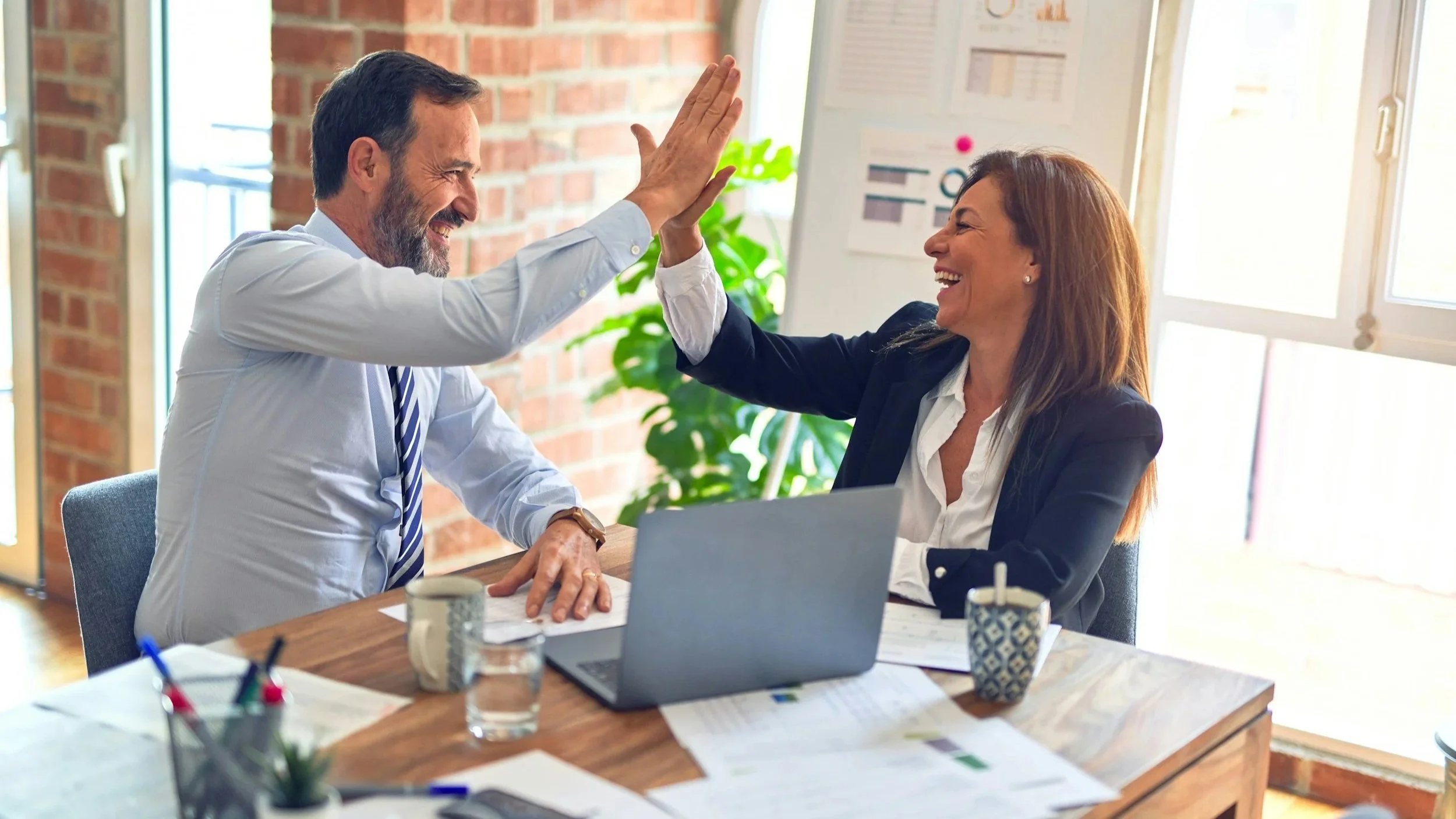 Man and woman high-fiving in an office, smiling, with documents, a laptop, and coffee mugs on the table.